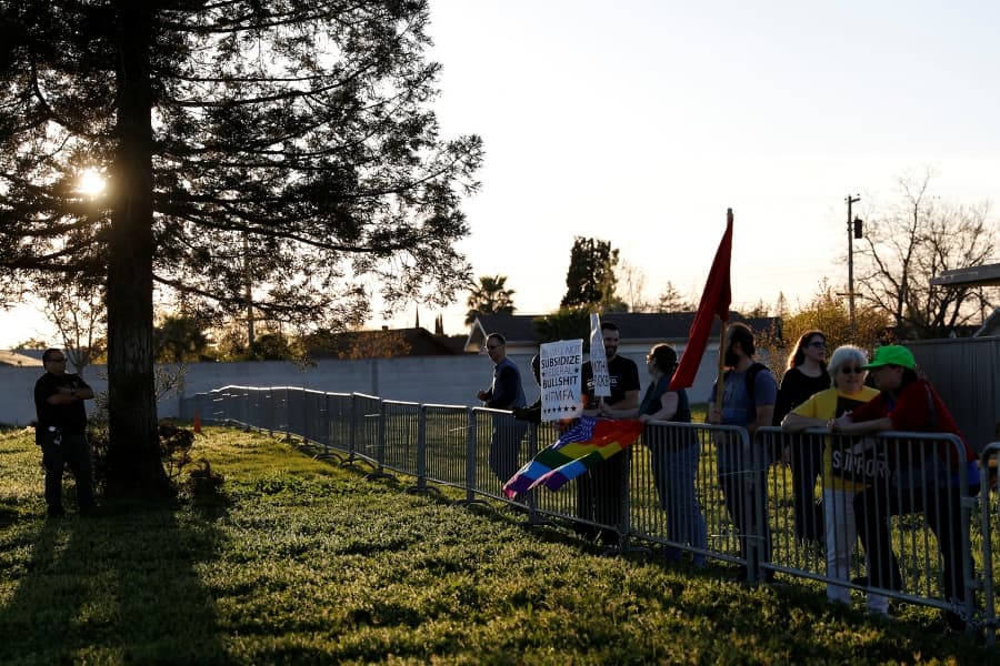 Line of protesters on lawn behind fence, with police officer near tree at edge of photo