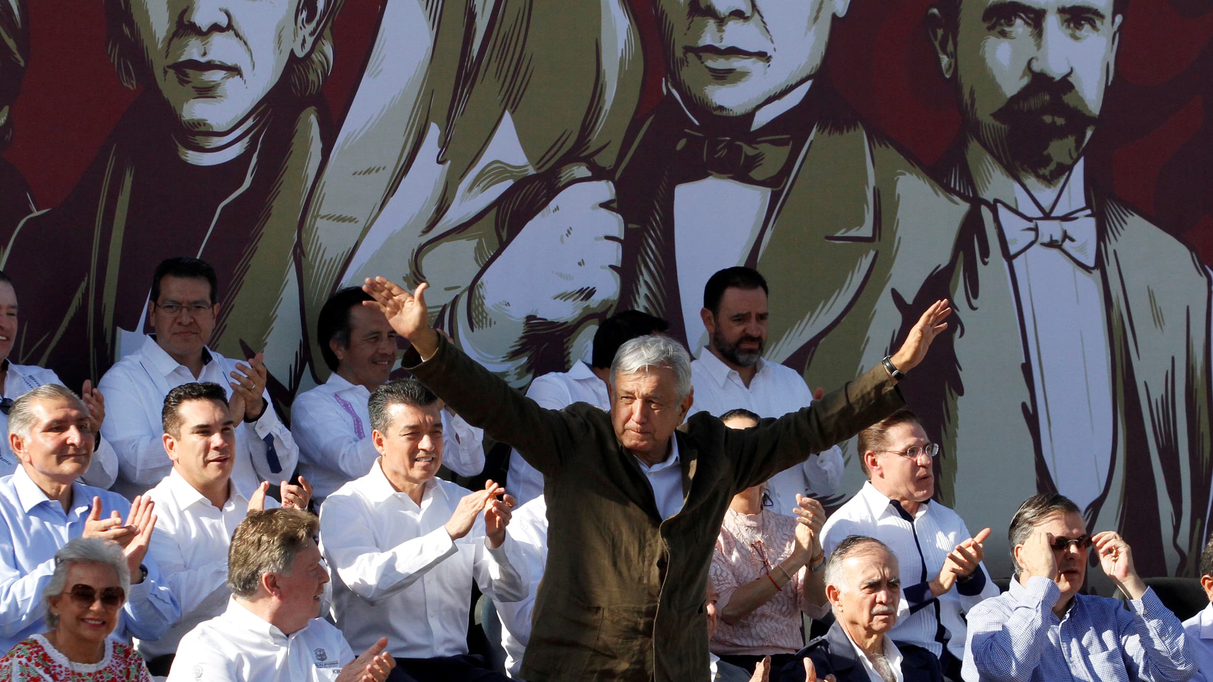 A man stands at a podium, his arms raised as he speaks. Others are seated around him.
