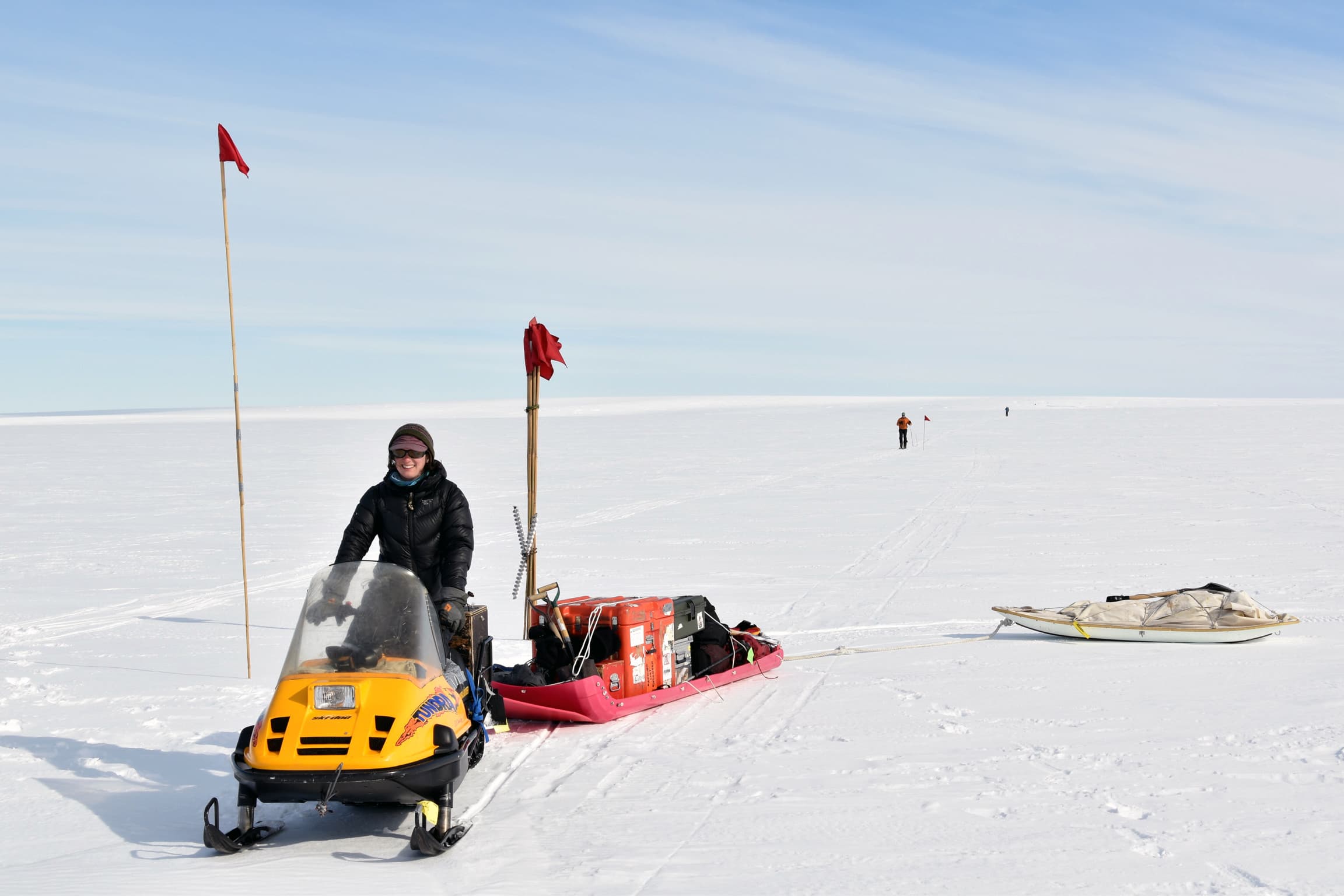 A woman steers a snowmobile towing equipment across the icy landscape.