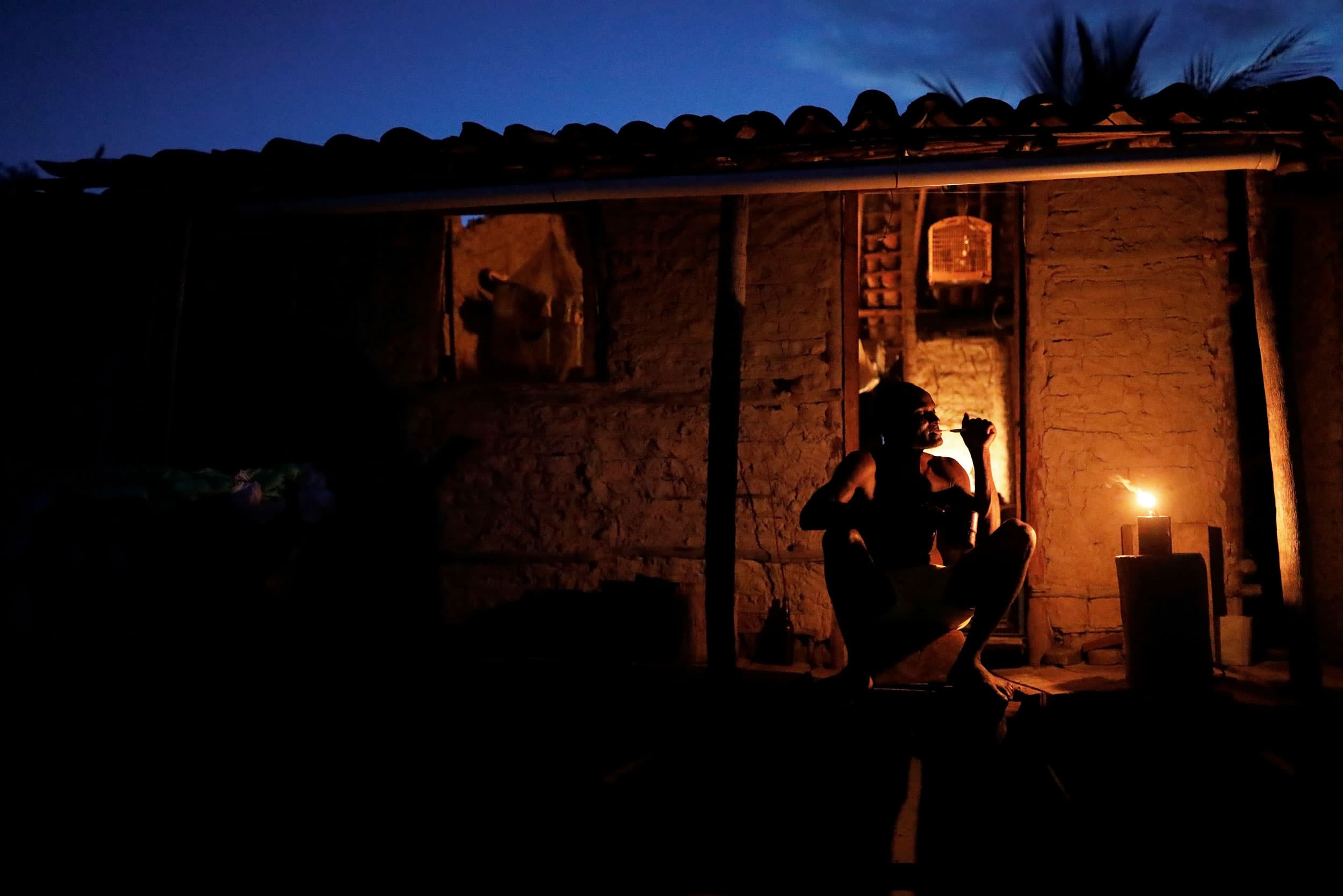 A man is shown sitting and brushing his teeth in a dark photograph at night with only the light from the house glowing.