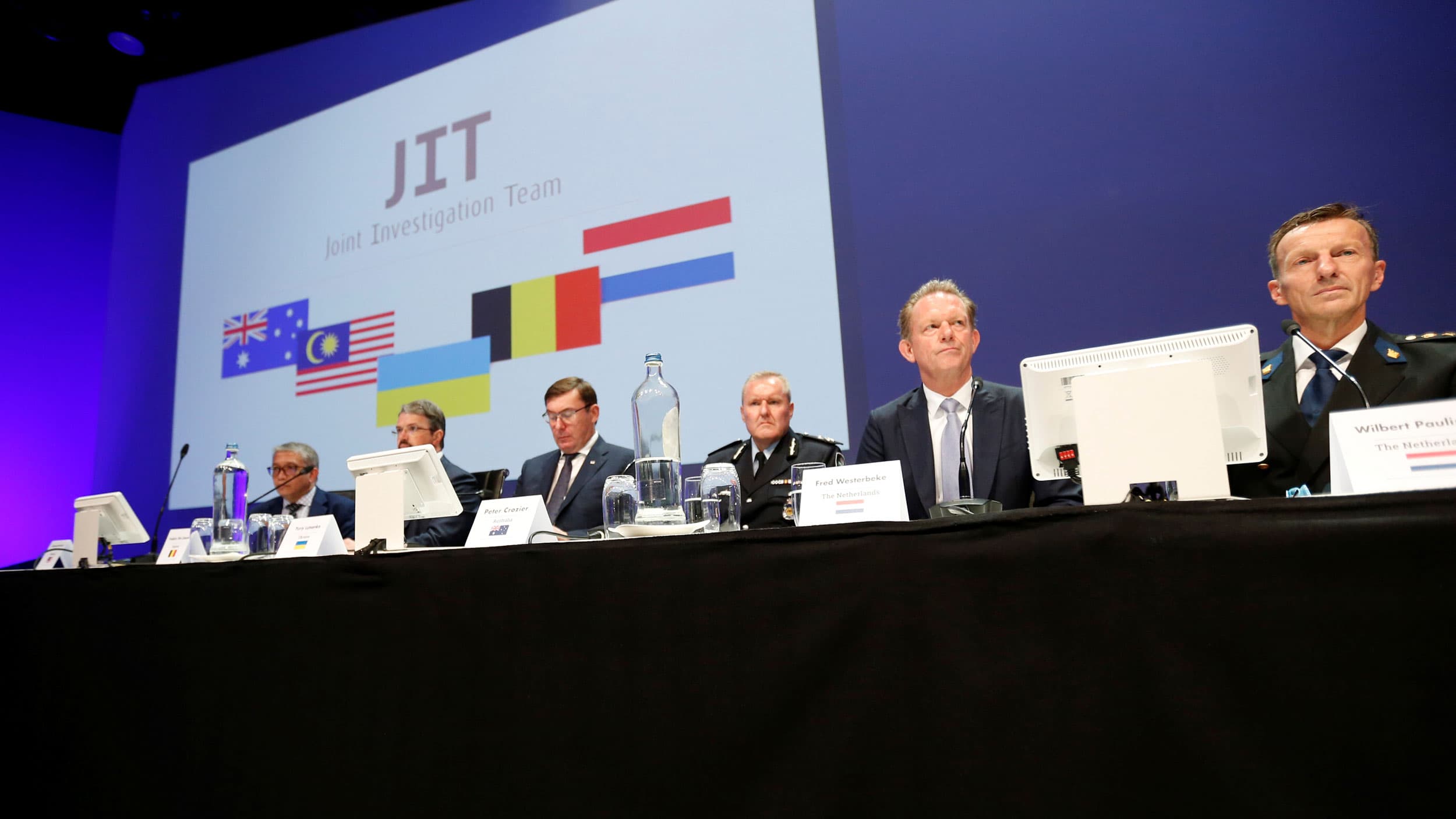 Six men are show sitting side-by-side at a table with a sign hanging behind them with "Joint Investigation Team" written on it.