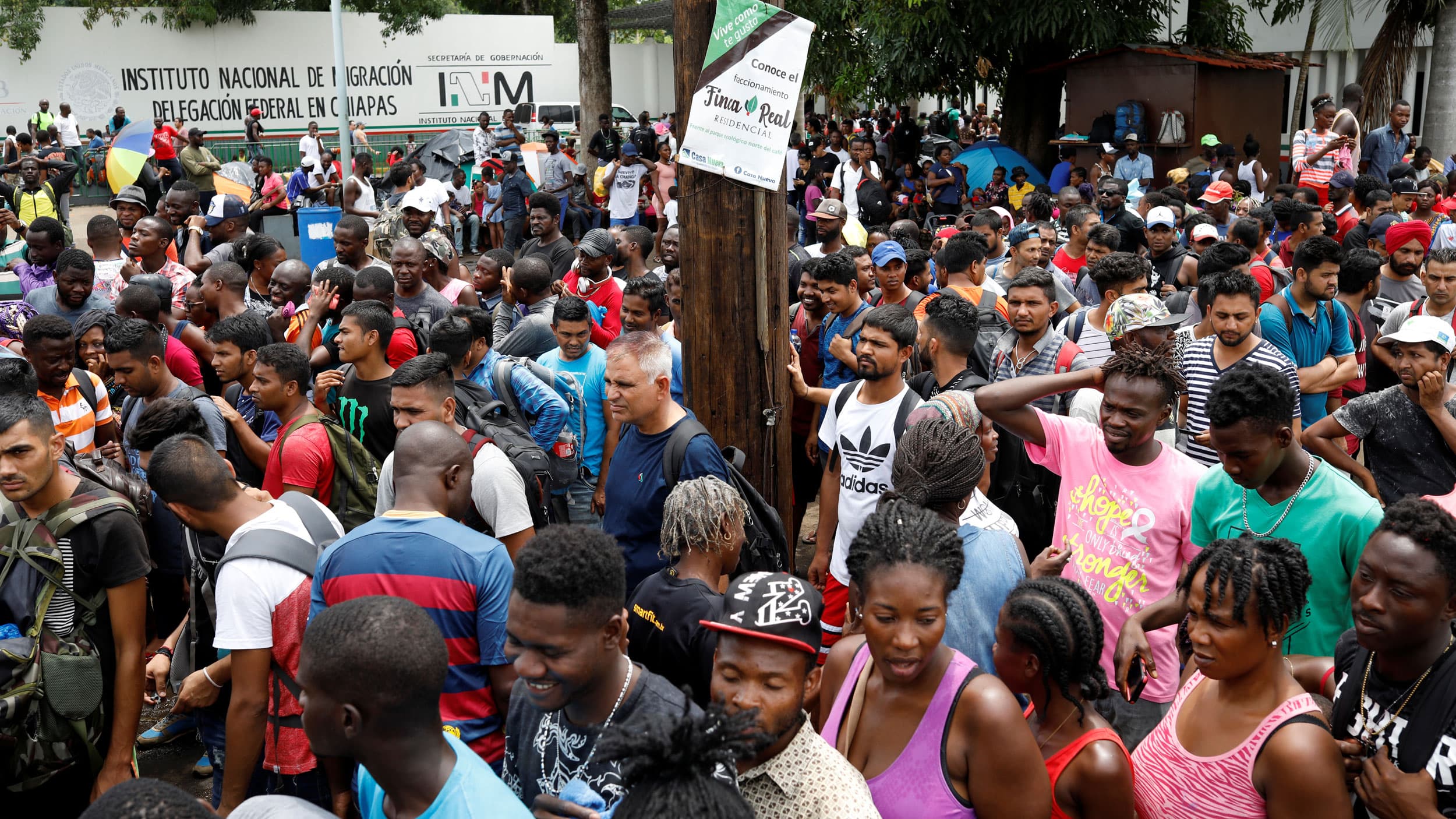 A large group of people are show standing outside of a white-walled building with the words "Instituto Nacional de Migracion" written on it.