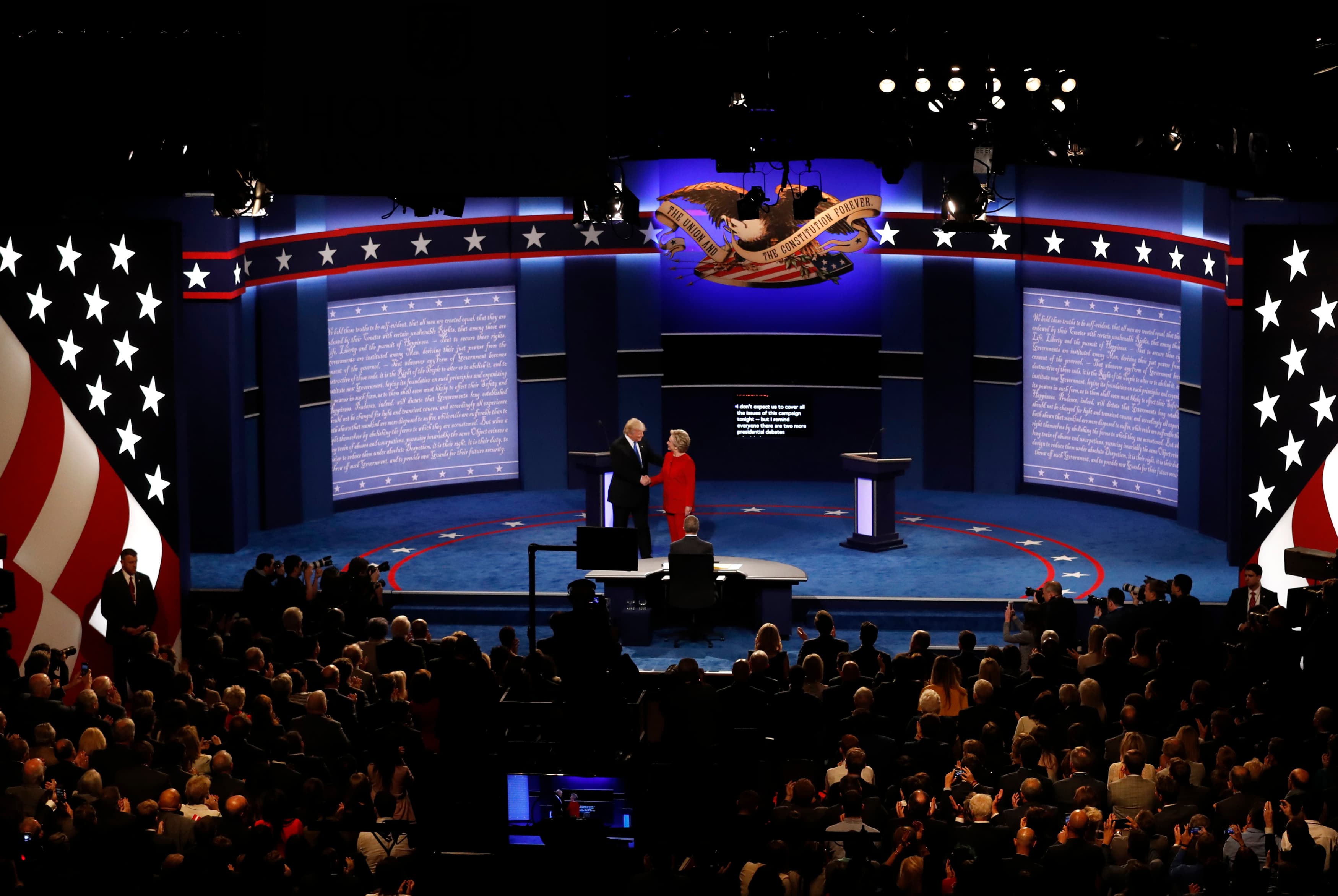Donald Trump and Hilary Clinton shake hands on stage in front of a crowd.