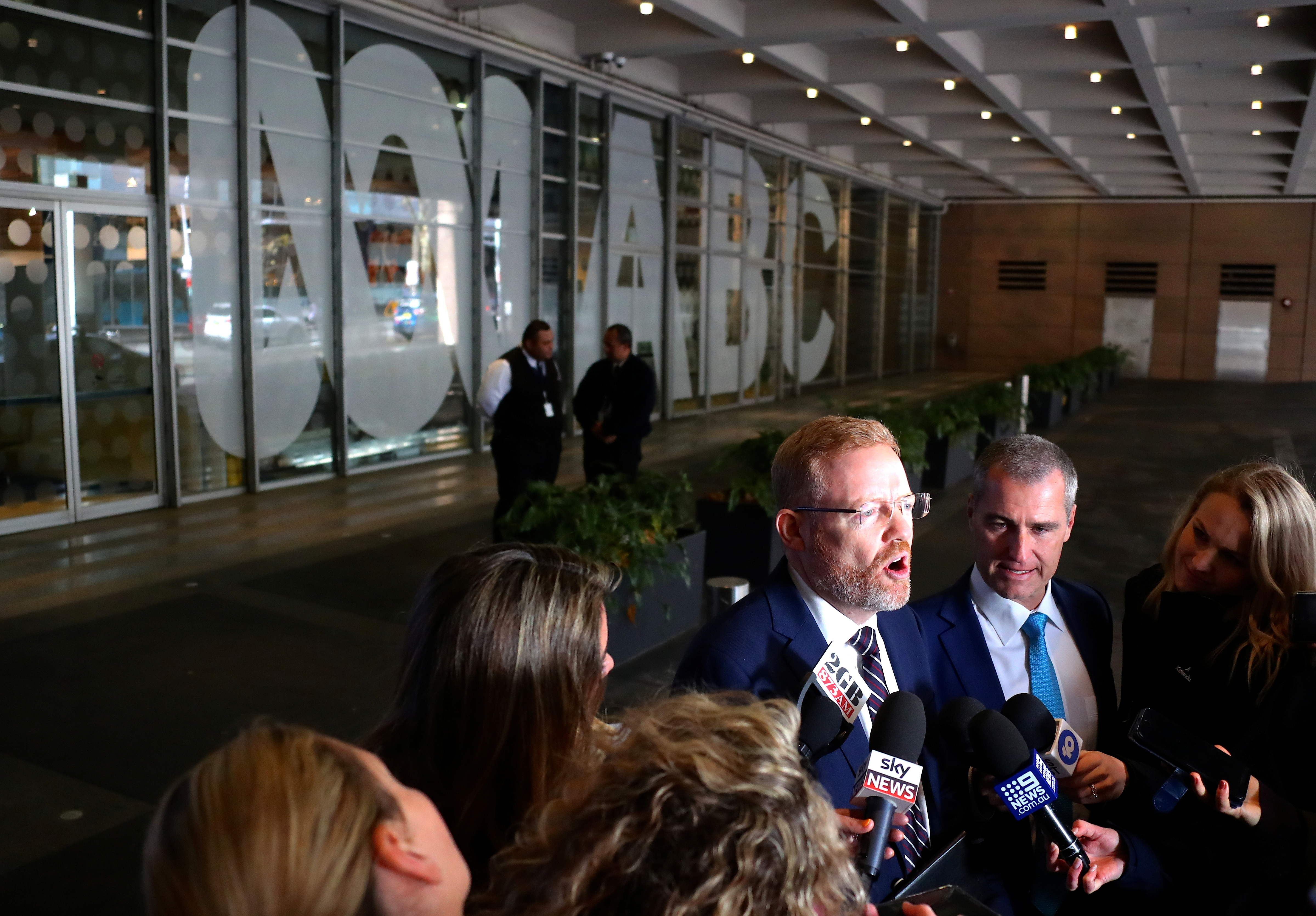 A man speaks in front of a crowd of journalists with microphones.