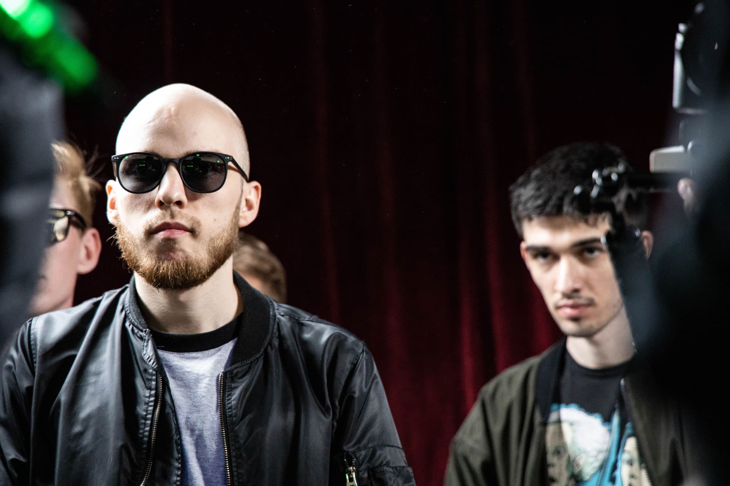 Two men stand next to each other and watch a rap battle.