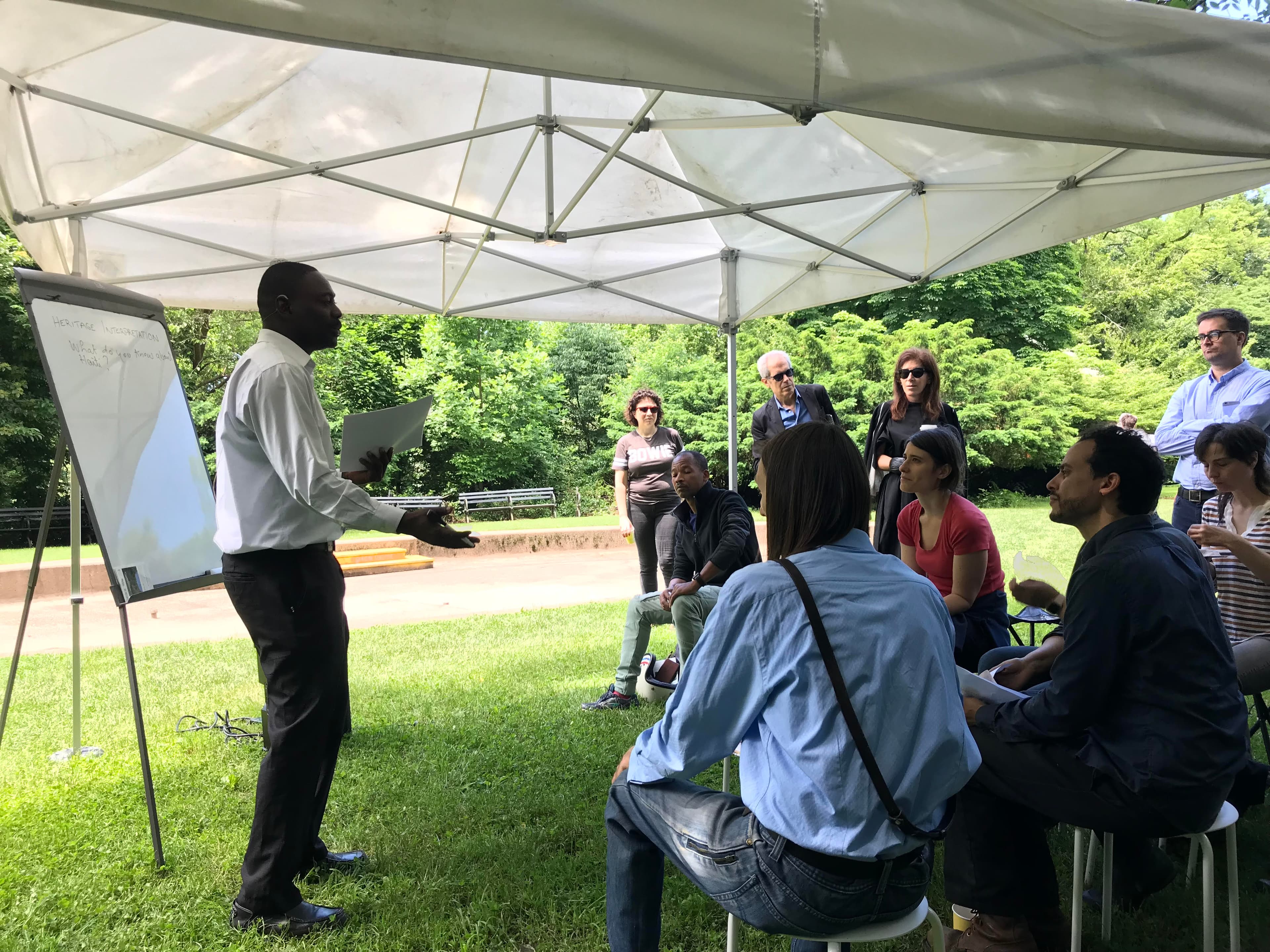 A man stands under a tent talking to people sitting down