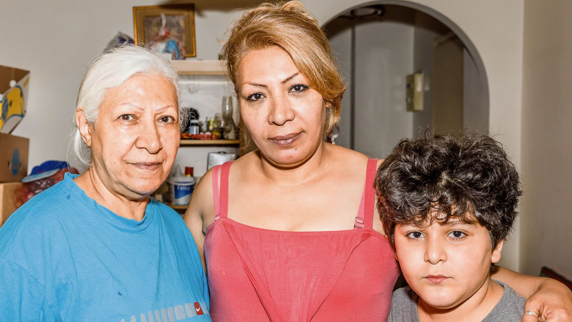 A grandmother wearing a blue shirt, stands to the left of her daughter wearing a red dress who is standing next to her son.