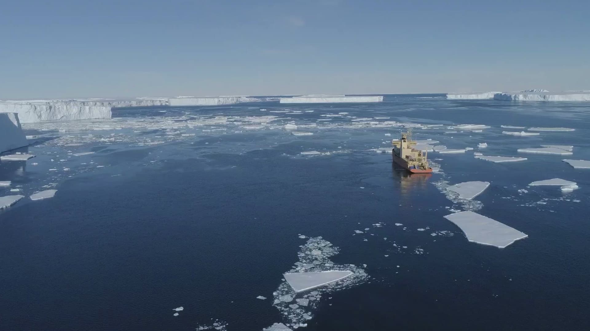 An overhead view of Antarctica. There are many white icebergs in a deep blue sea and a bright orange ship