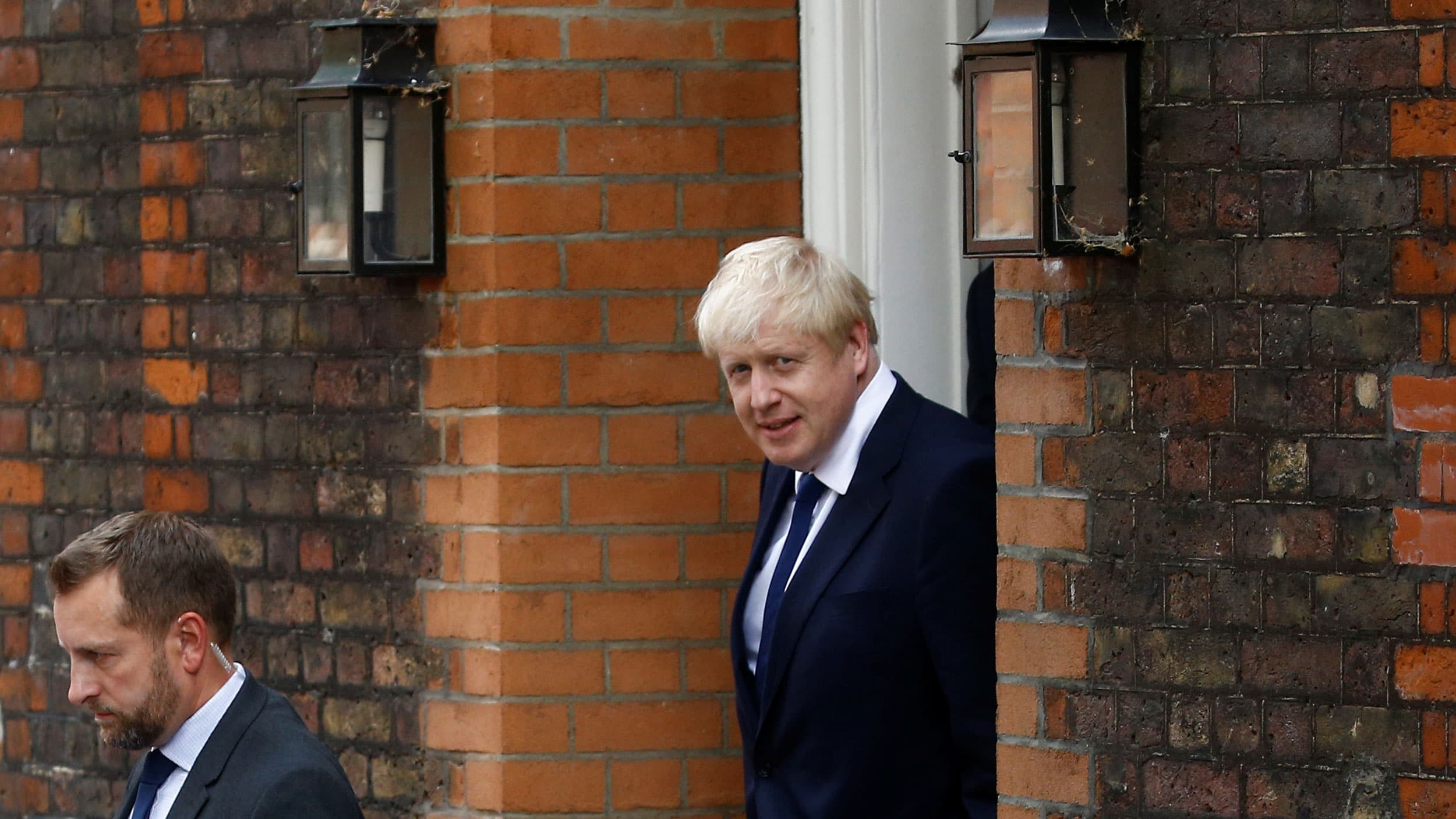 Boris Johnson is shown walking through a brick doorway wearing a blue suit.