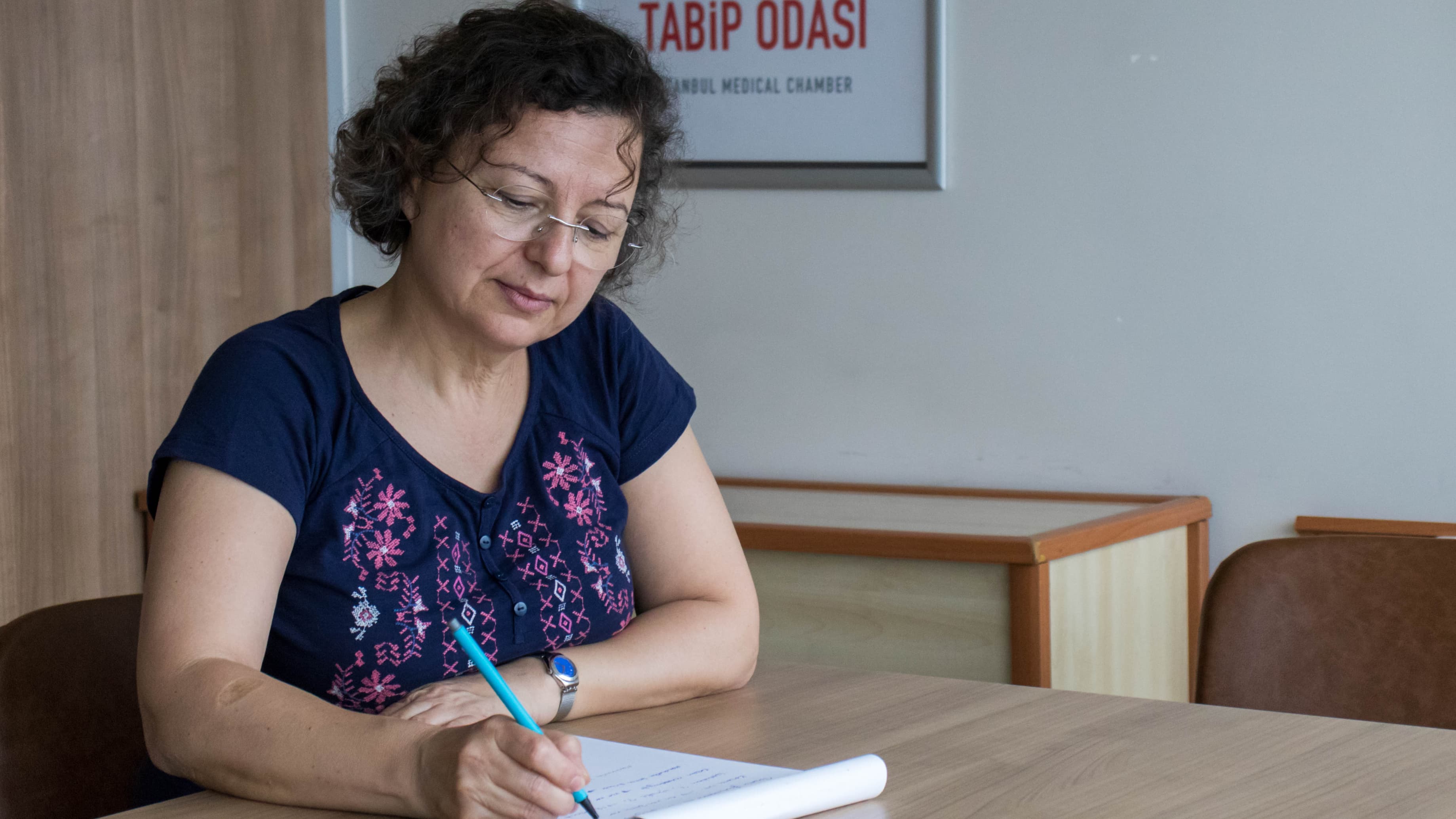 A woman sits at a table and writes on a notepad.