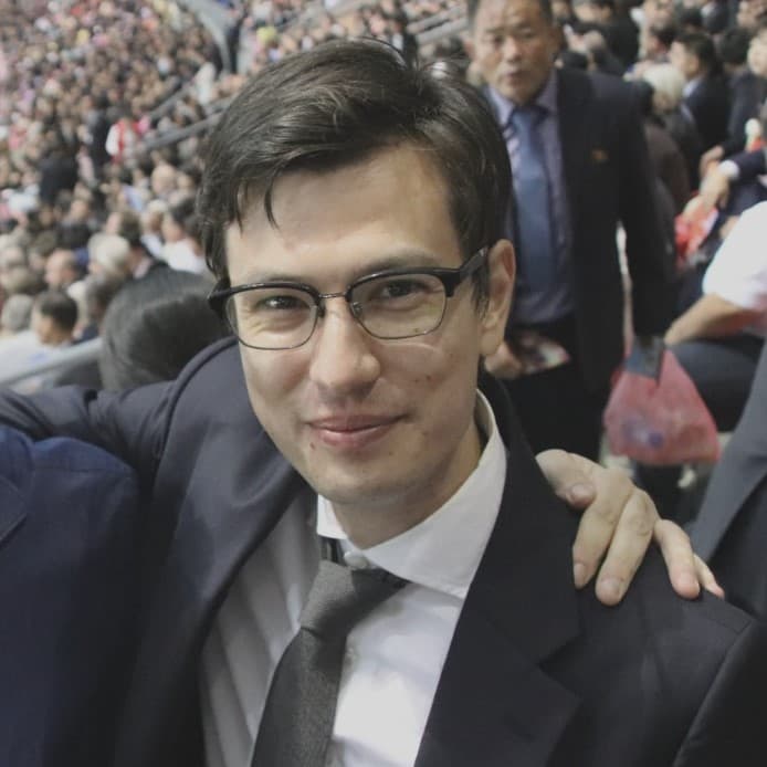 A close up of Alex Sigley in glasses and a suit with a dark tie.