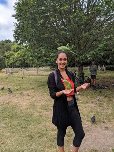 a woman holds a parakeet as another one sits on her head
