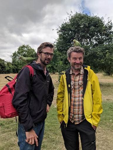 two men have parakeets on their heads in a london park