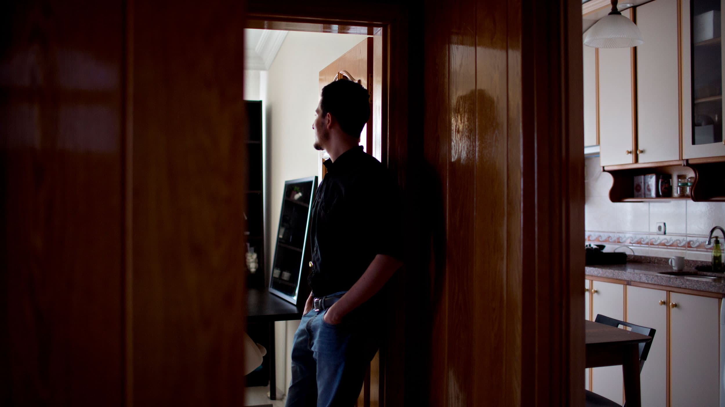 A man stands in a doorway looking off with a kitchen shown around the corner.