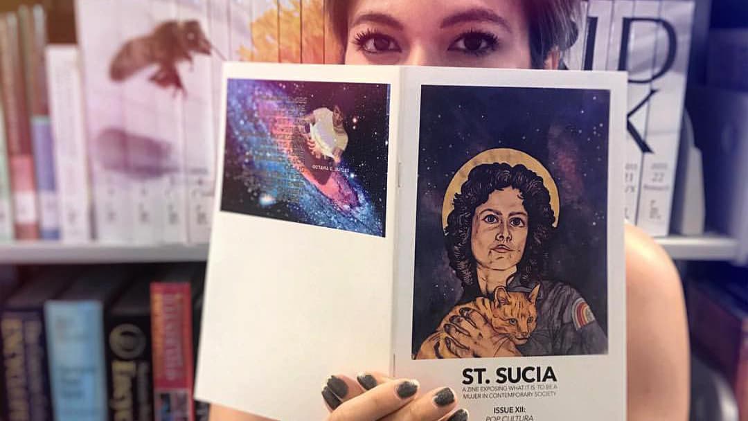 A woman holds open a magazine with an illustration of an astronaut and cat on the cover. The woman's eyes are visible above the pages.