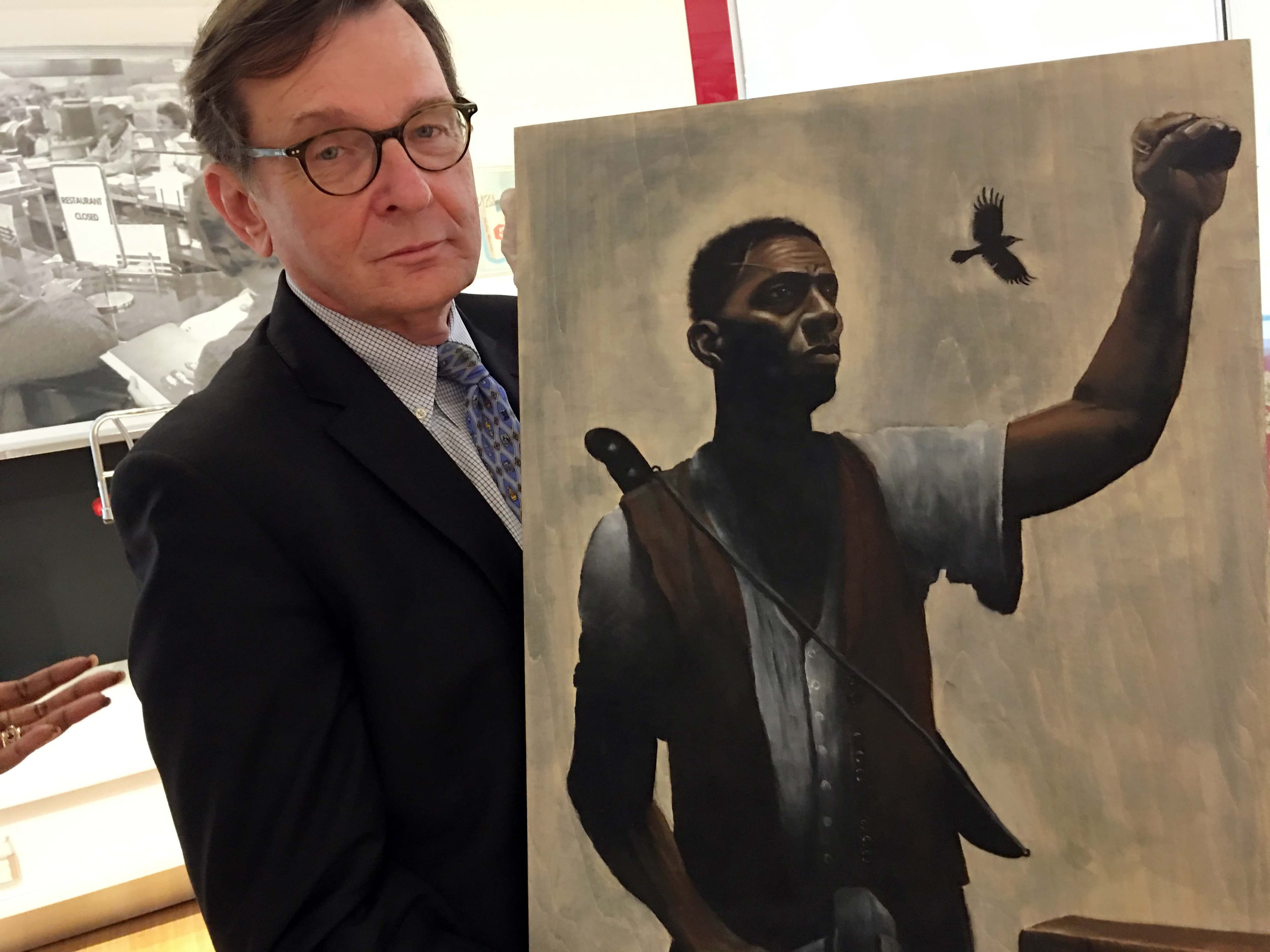 A white man in glasses and a suit holds a painting of a black man with a raised fist wearing a shirt and vest.