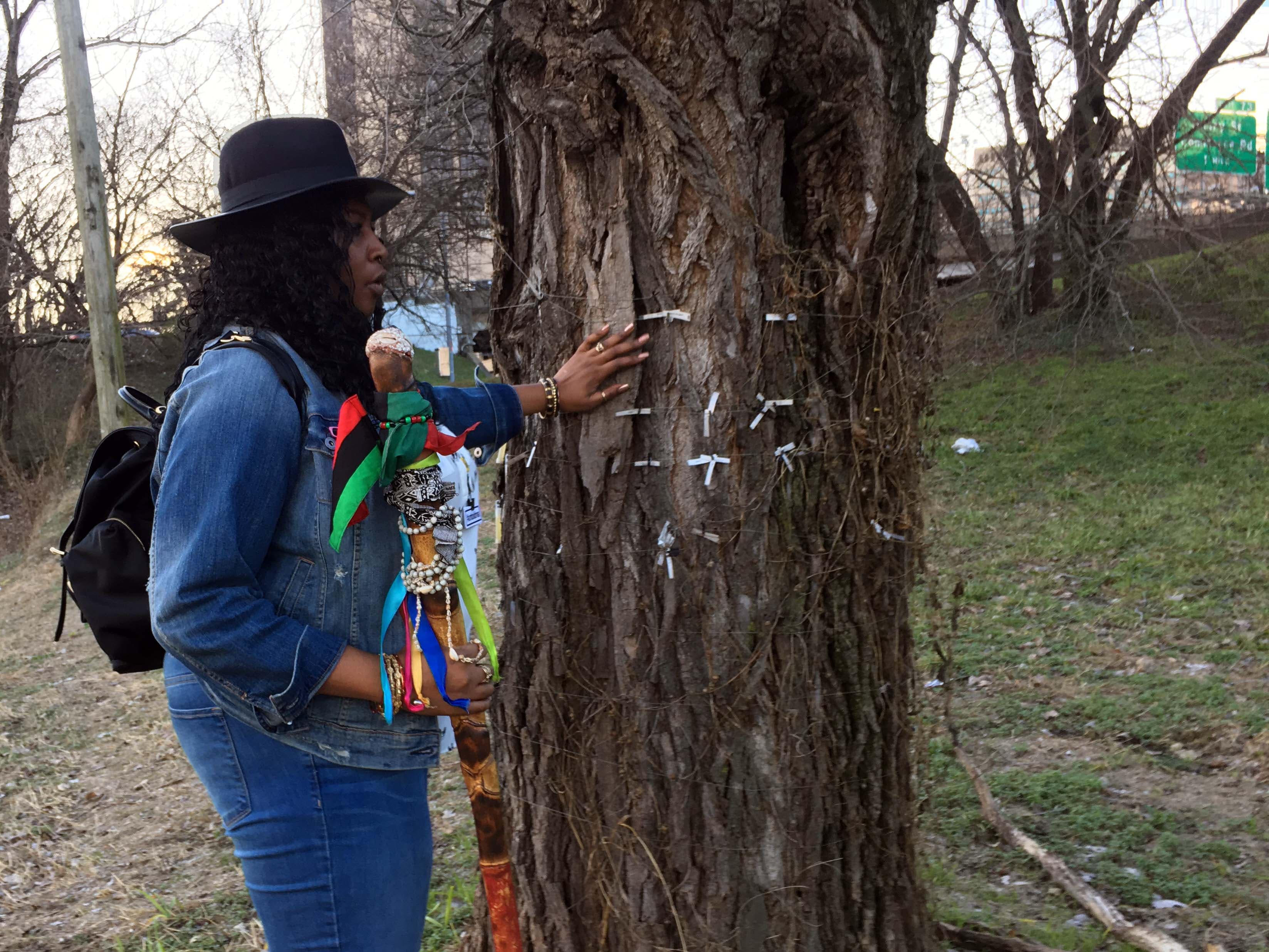 A black woman in a hat stands next to a tree with tags hanging from it.