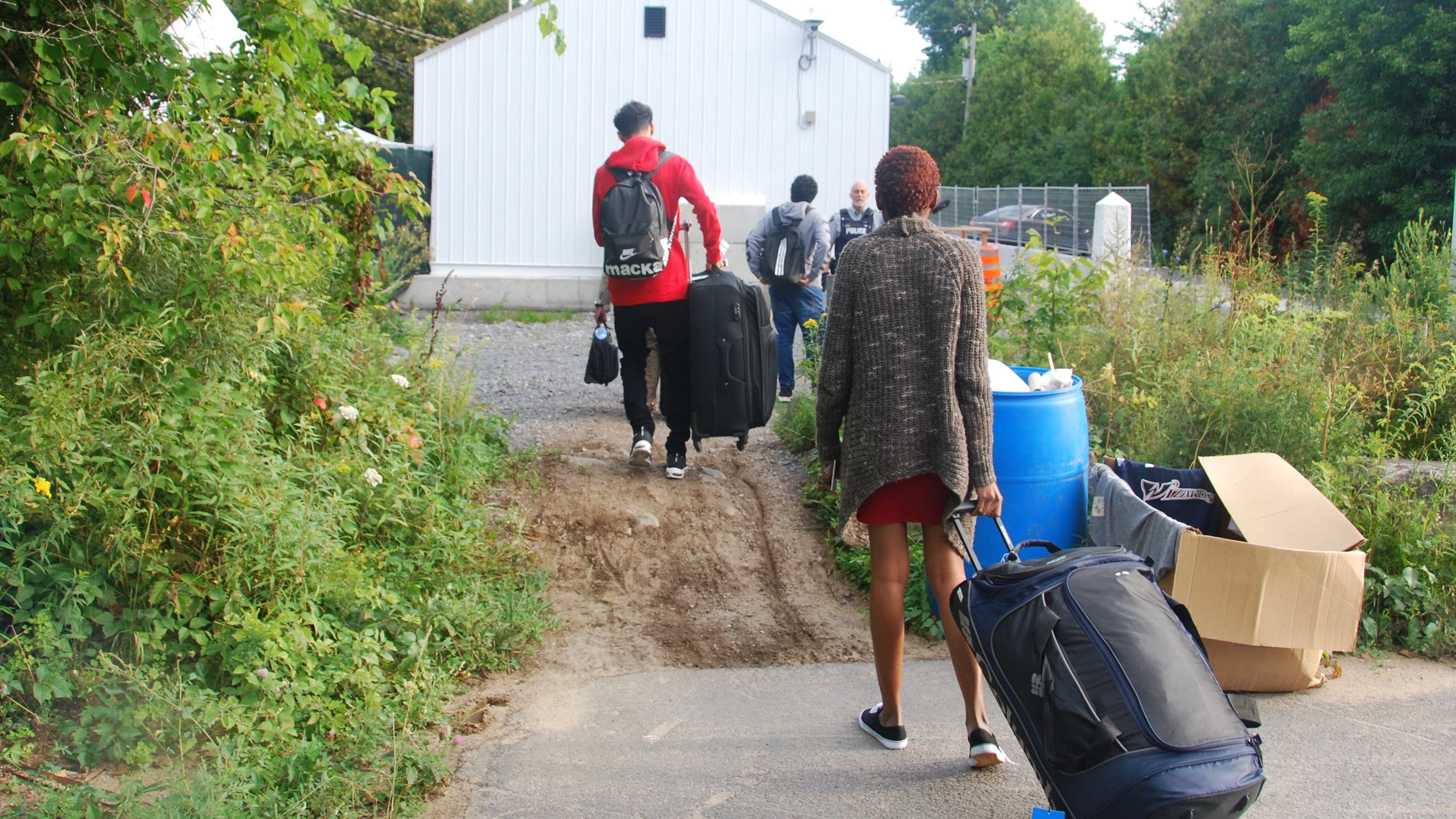 A woman drags a suitcase in the foreground. In the background, a police officer and three men dragging suitcases.