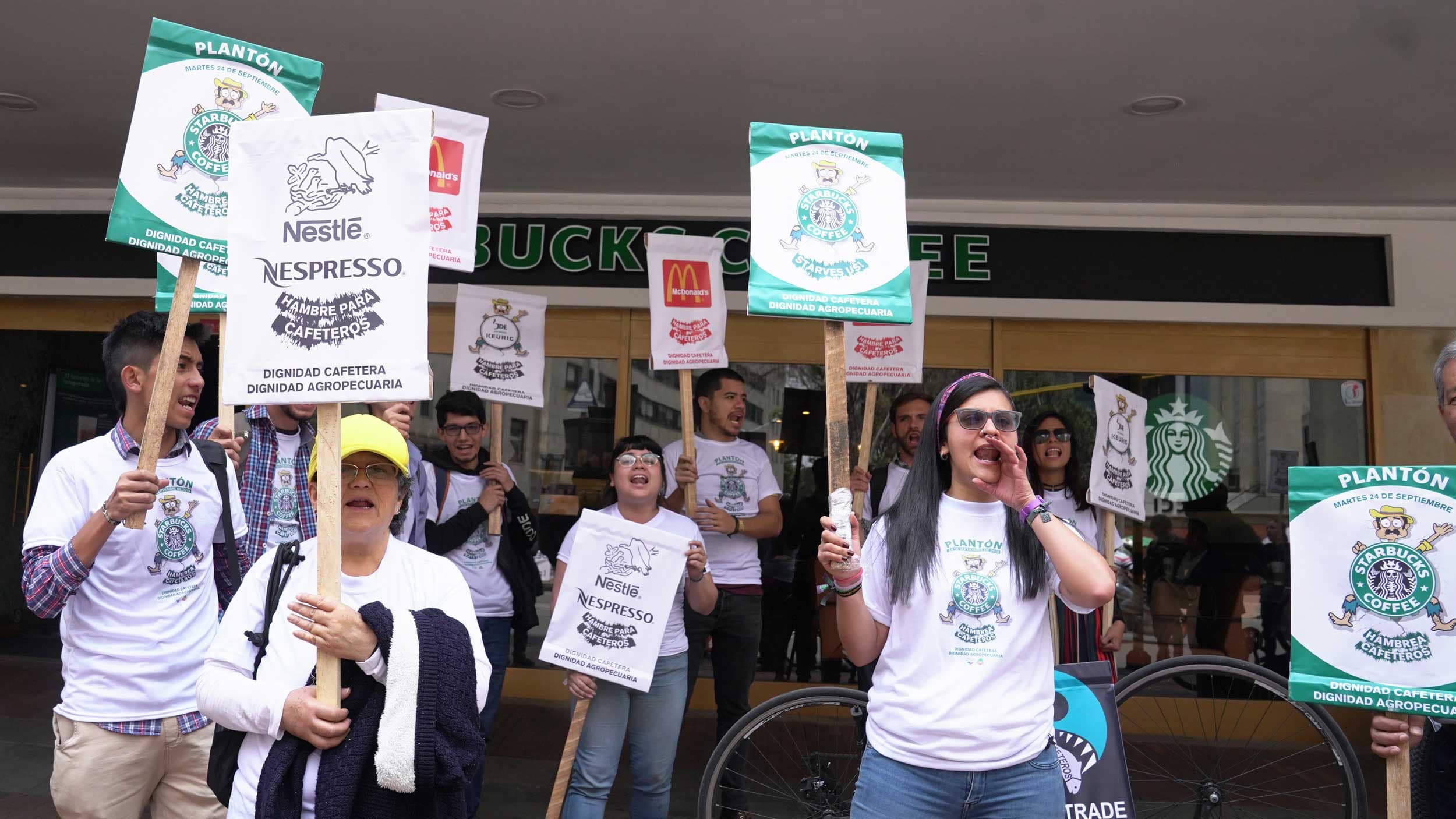 Protesters with signs stand in front of a Starbucks storefront.