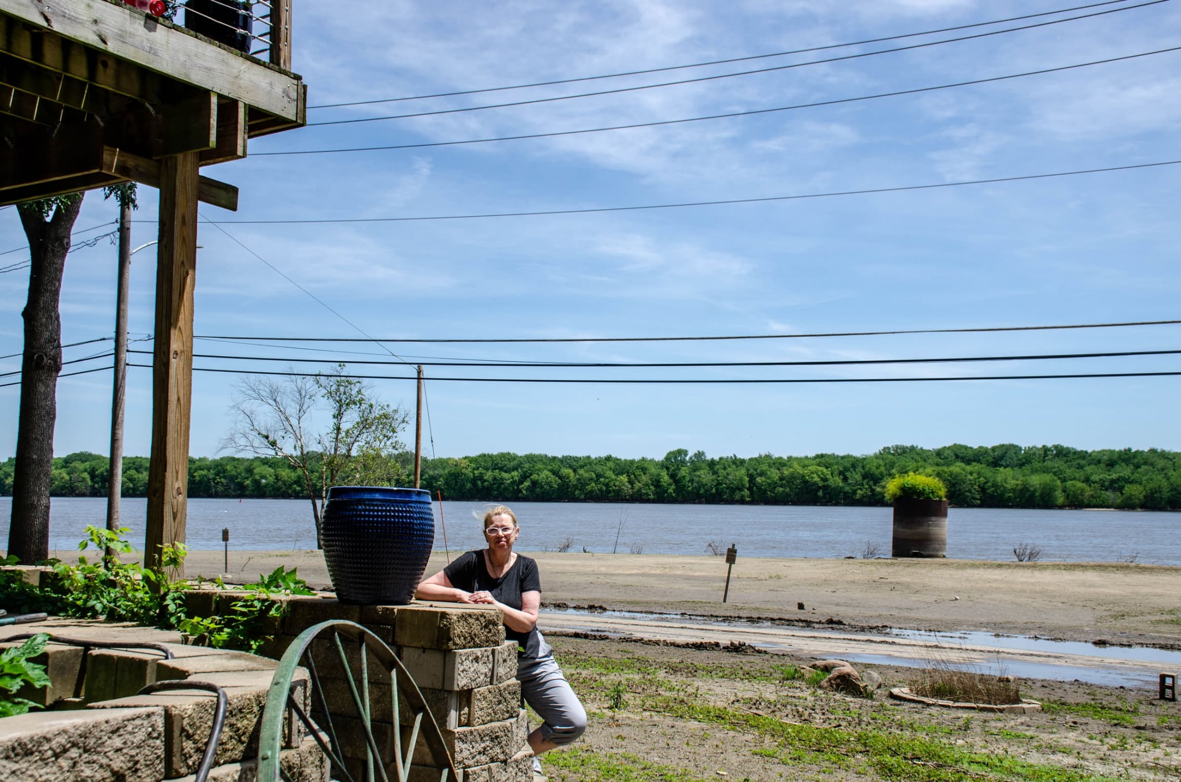 A woman is shown standing next to the base wall of her house which has been raised up on wooden beams.