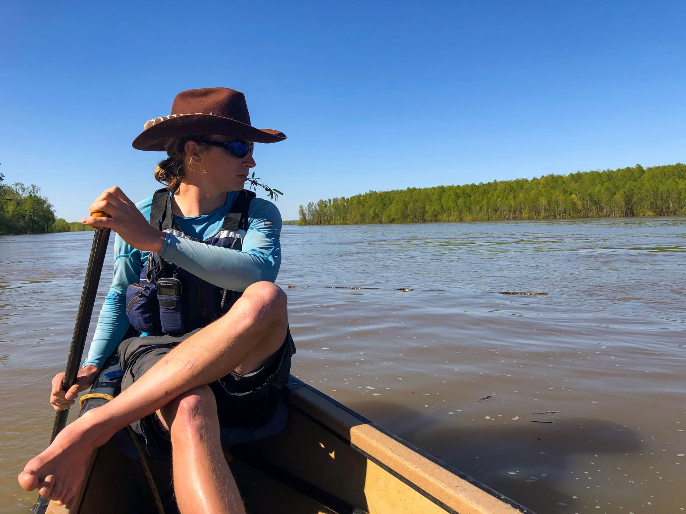 A man is shown in the back of a canoe with a paddle in his hand while wearing a wide-brimmed hat.