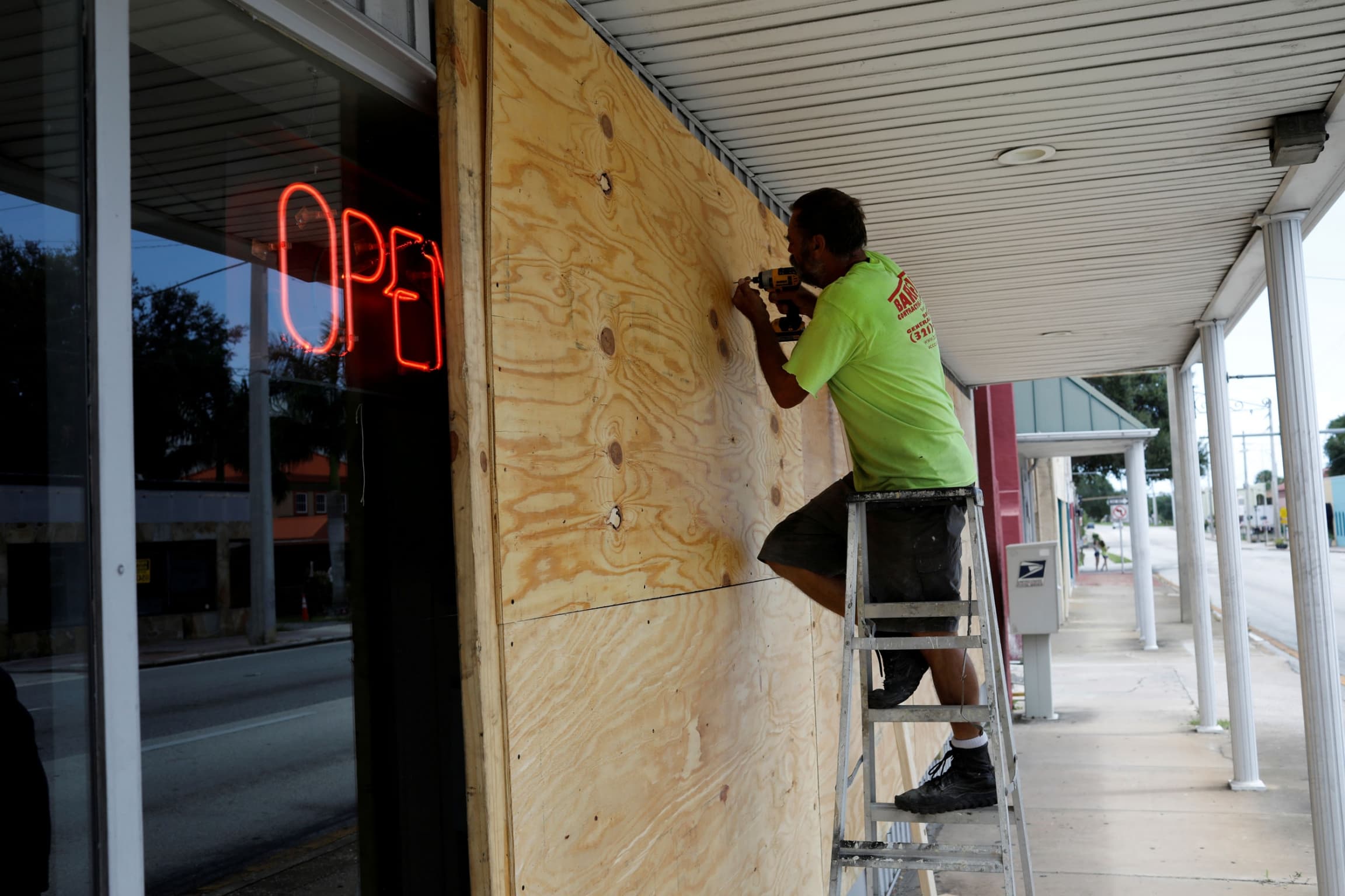 A man is shown standing on a ladder with a power drill in his hand fastening a wooden board to the front of a building.