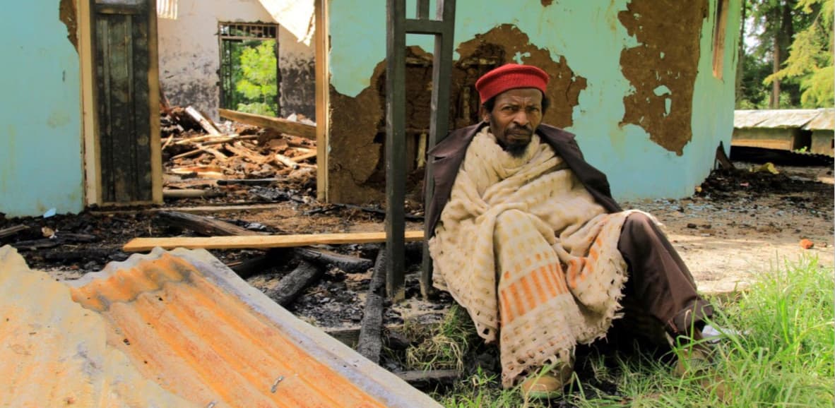 A man with a red cap sits in front of a destroyed building.