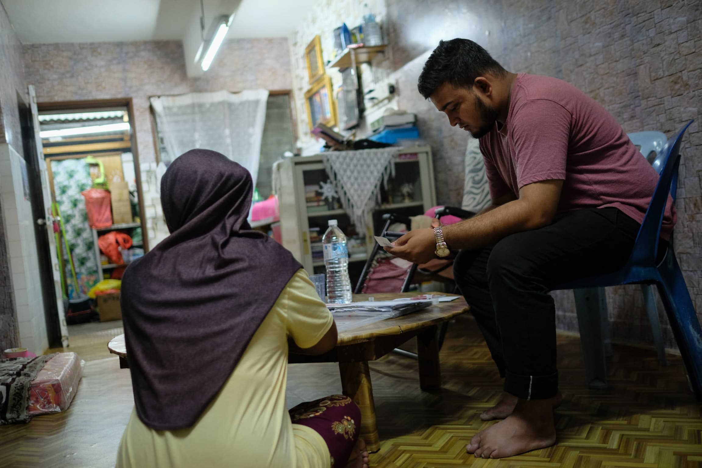 A young man sits on a stool near a woman wearing a headscarf with back toward the camera