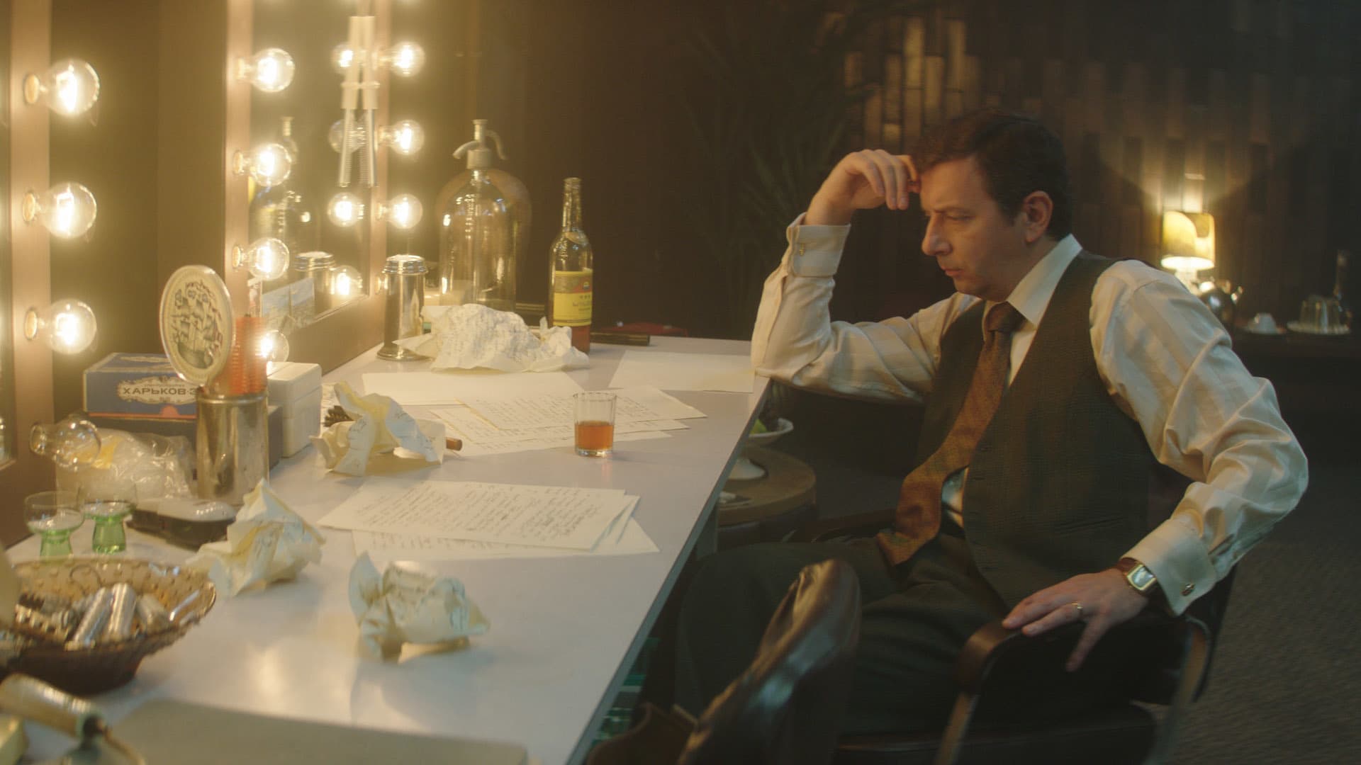 A film still of a man sitting at a dressing room mirror with lights