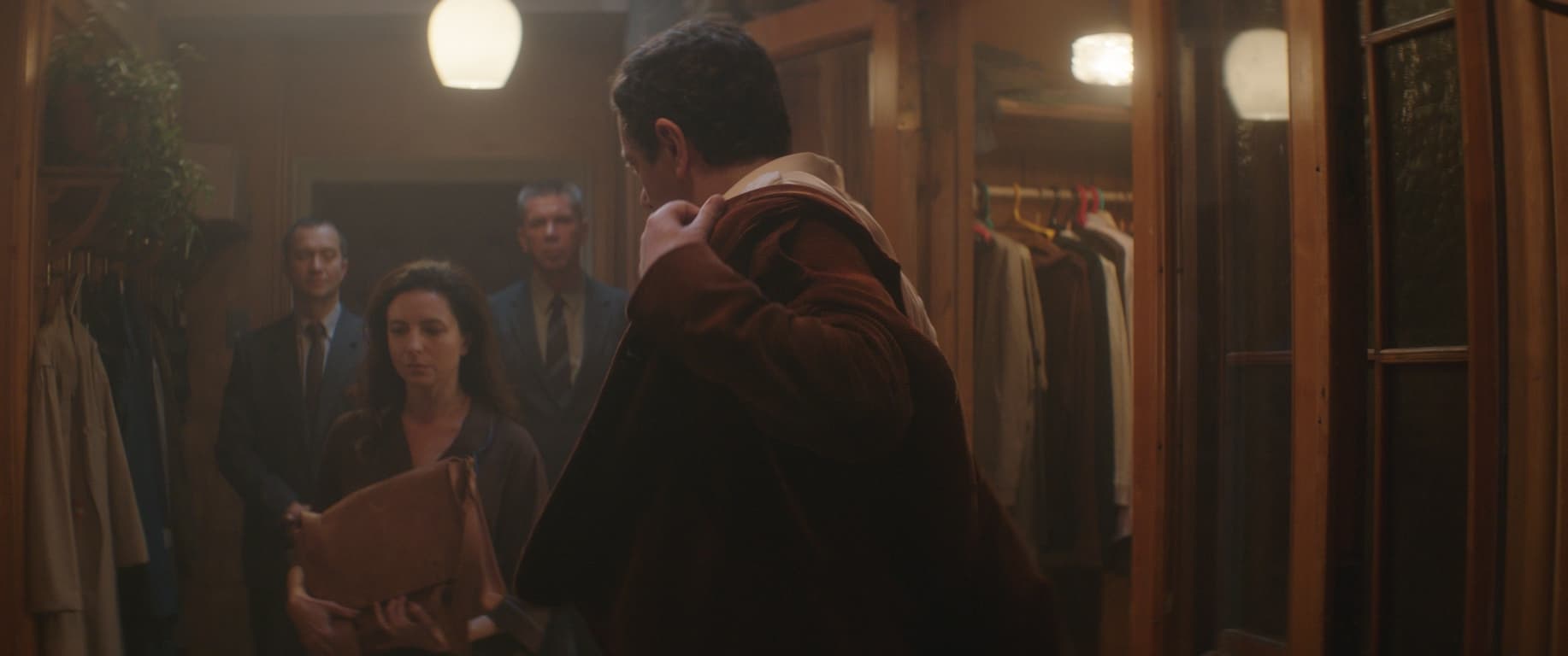 A sepia-toned film still, two men in suits stand in the background, a woman in the midground is upset, a man in the foreground puts on his coat.