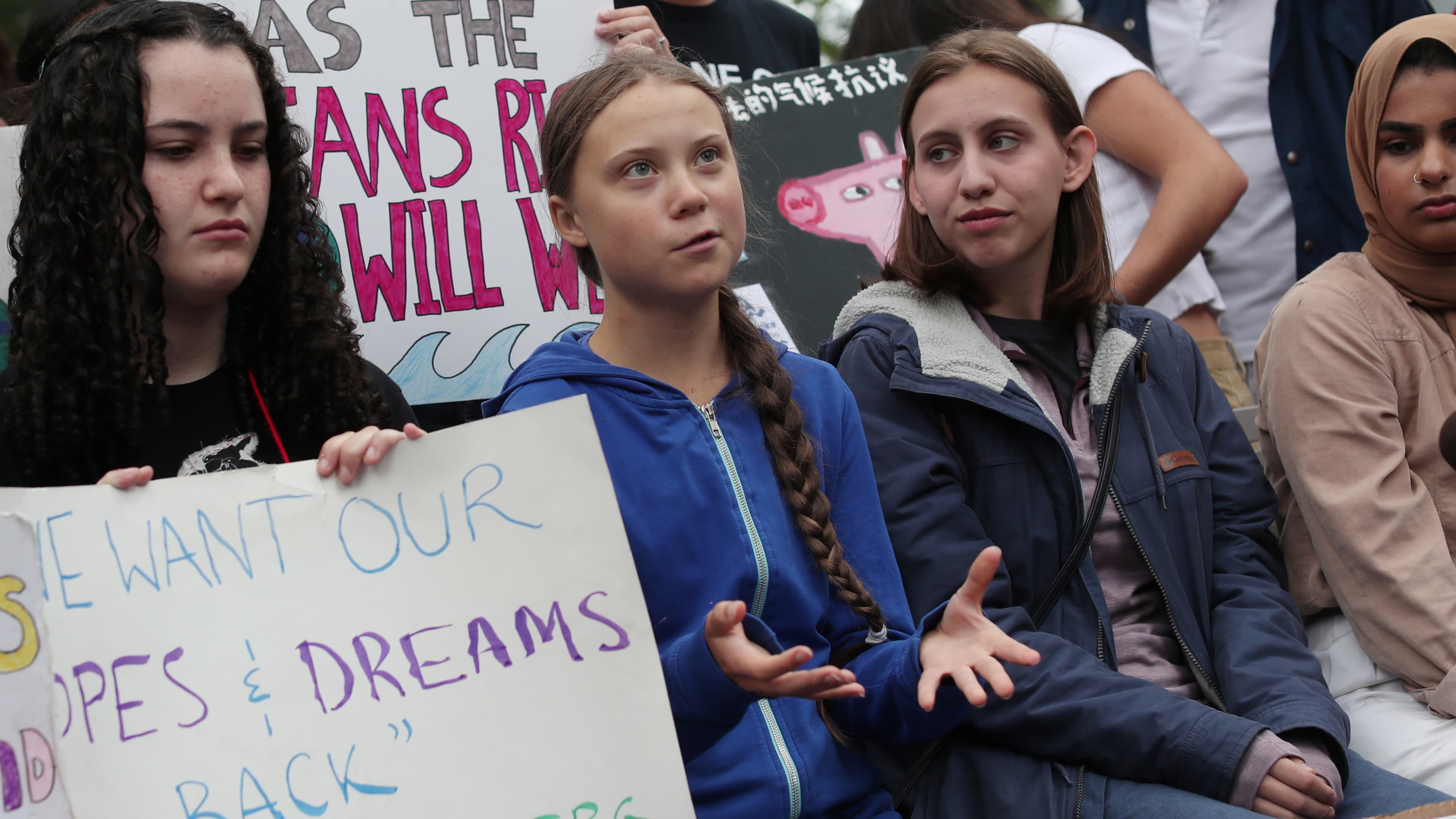 Four girls sit in a row of protesters, one with a long braid is speaking and gesturing with her hands.