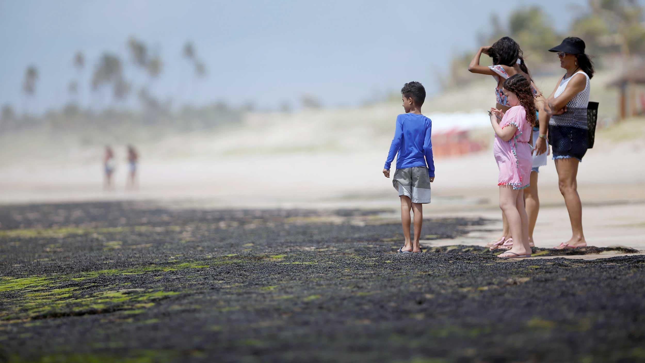 People stand on the sand of a beach. Below them, the sand is covered in a dark, black substance.