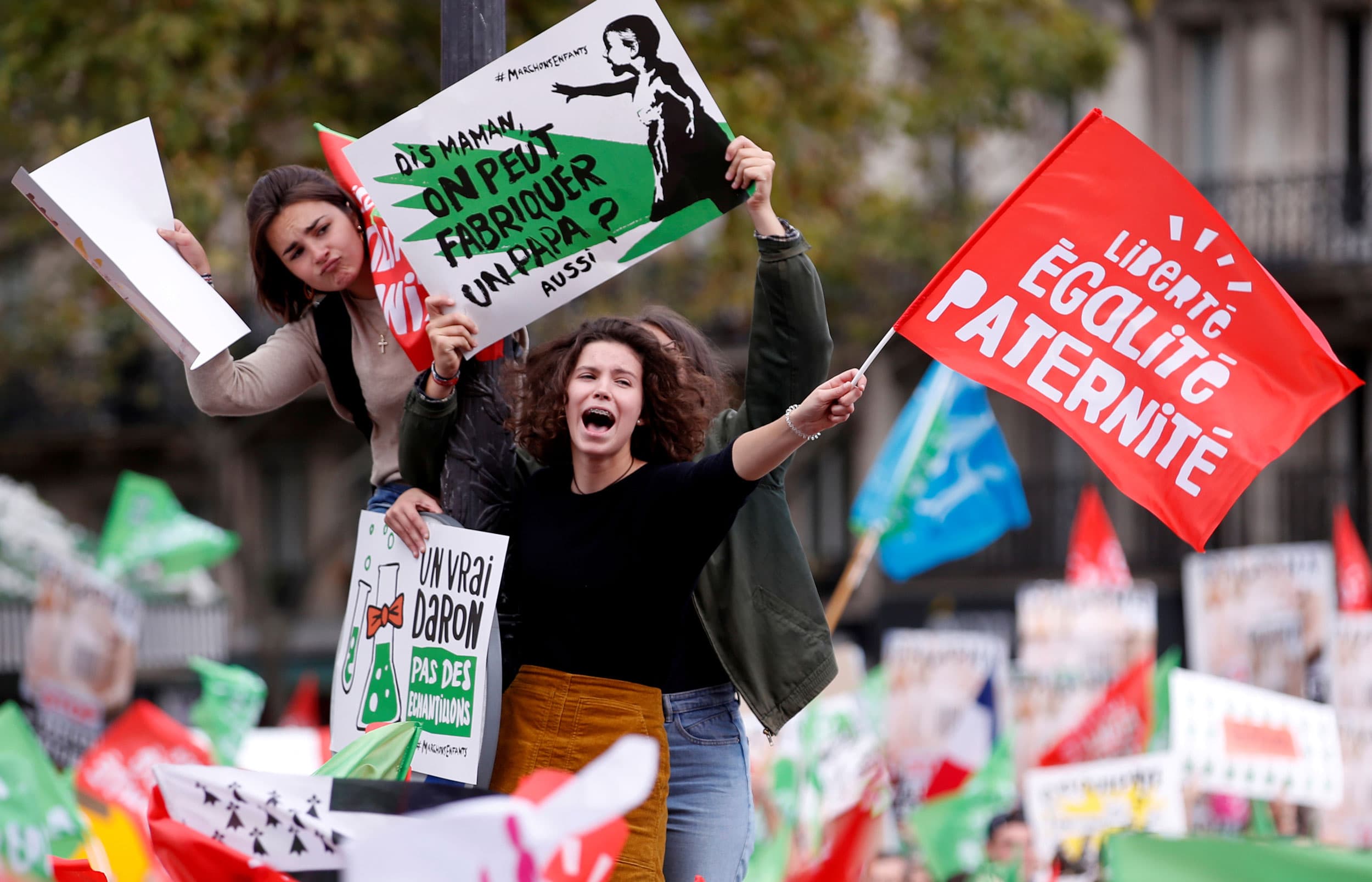 Woman hang off a street light carrying signs protesting a proposed bill. One of the signs is a play on the French motto: Liberty, equality, paternity!