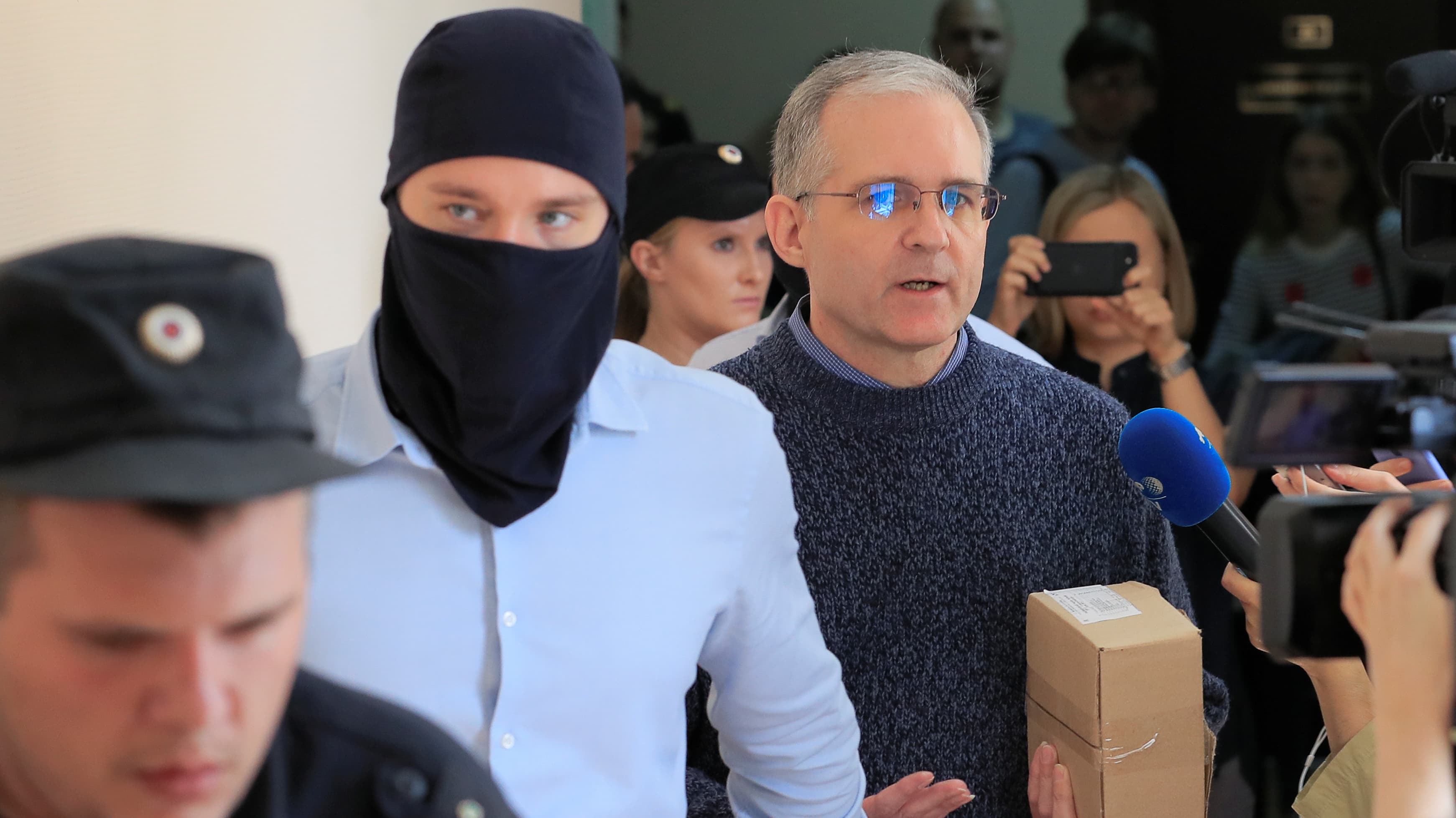 Three men walk into a court room, one is wearing a black mask, the other is holding a brown cardboard box.