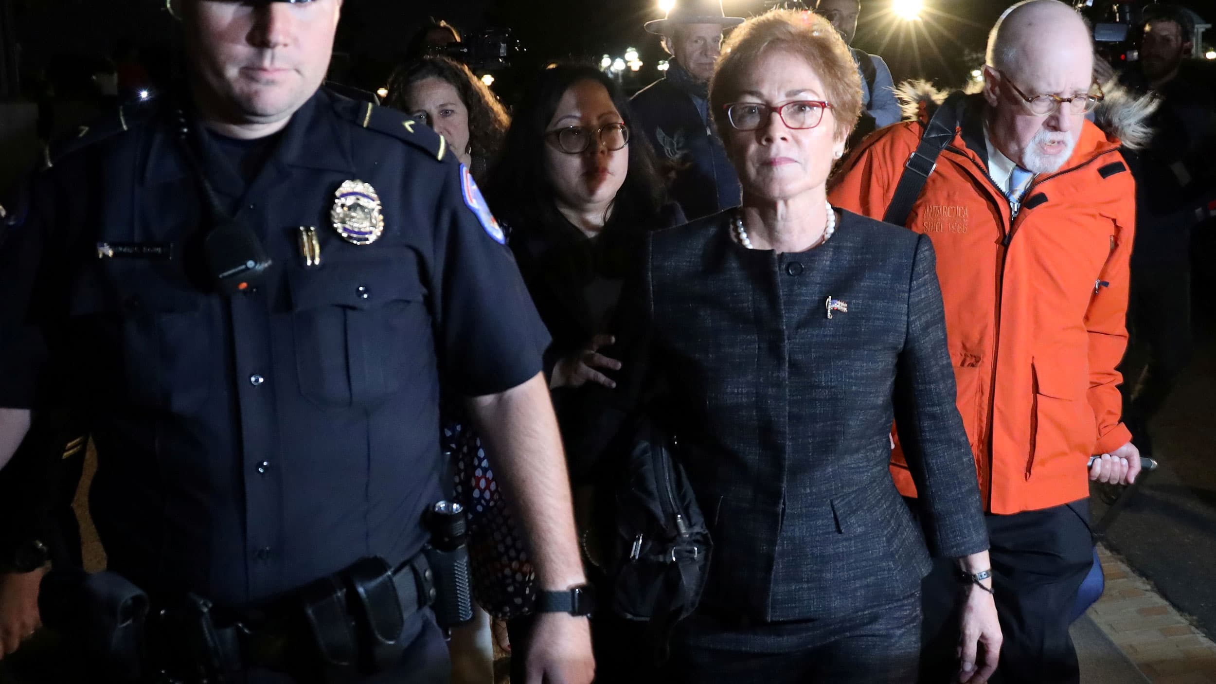 Police escort a woman wearing glasses. More people follow her.