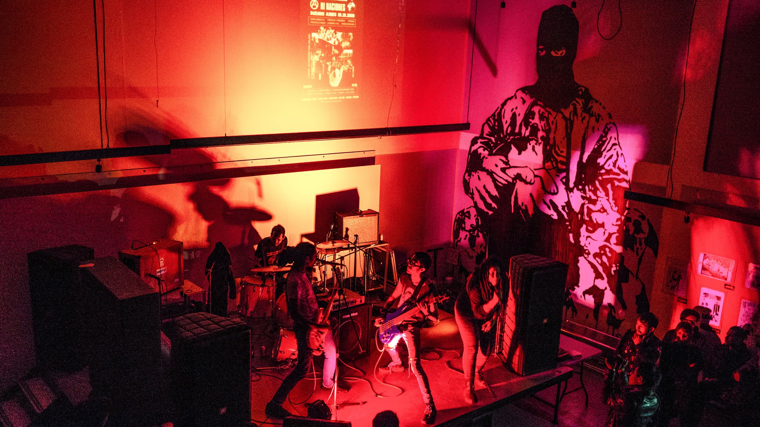 A four-piece punk band is shown playing to a crowd and bathed in red lights.