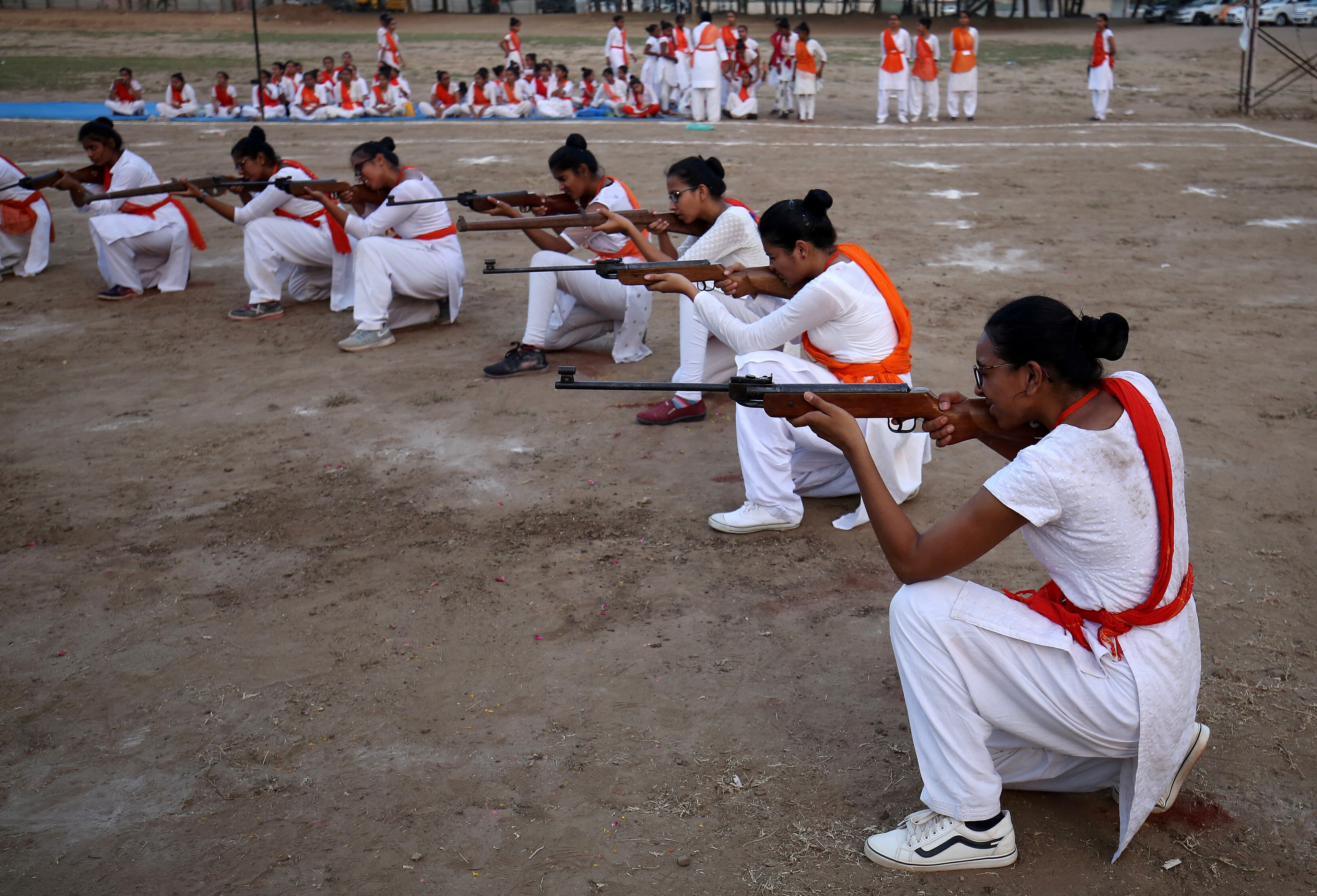 Women wear white with red sashes and hold guns aimed at a target.
