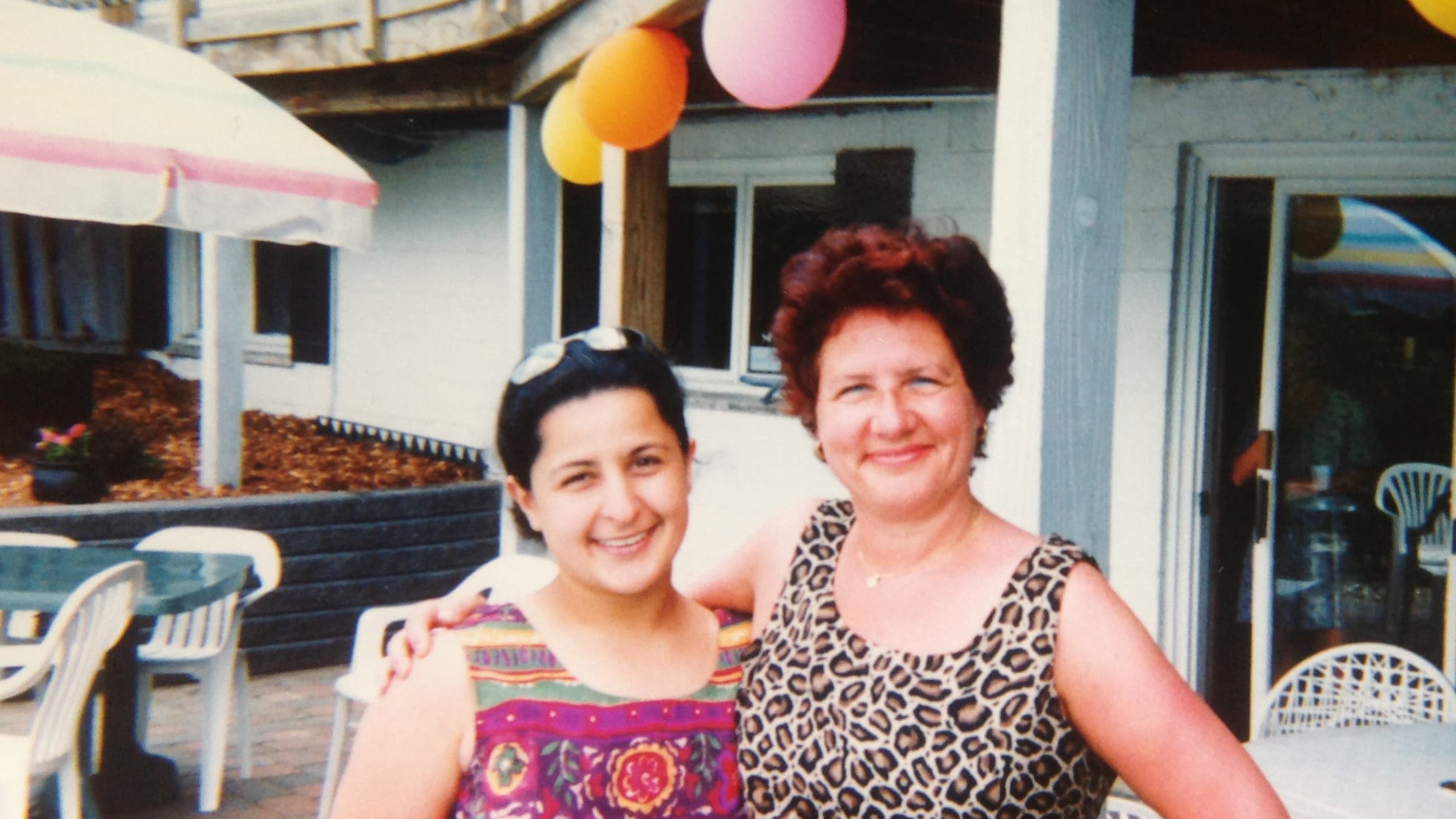 A young woman stands next to her mother with colorful balloons in a backyard.