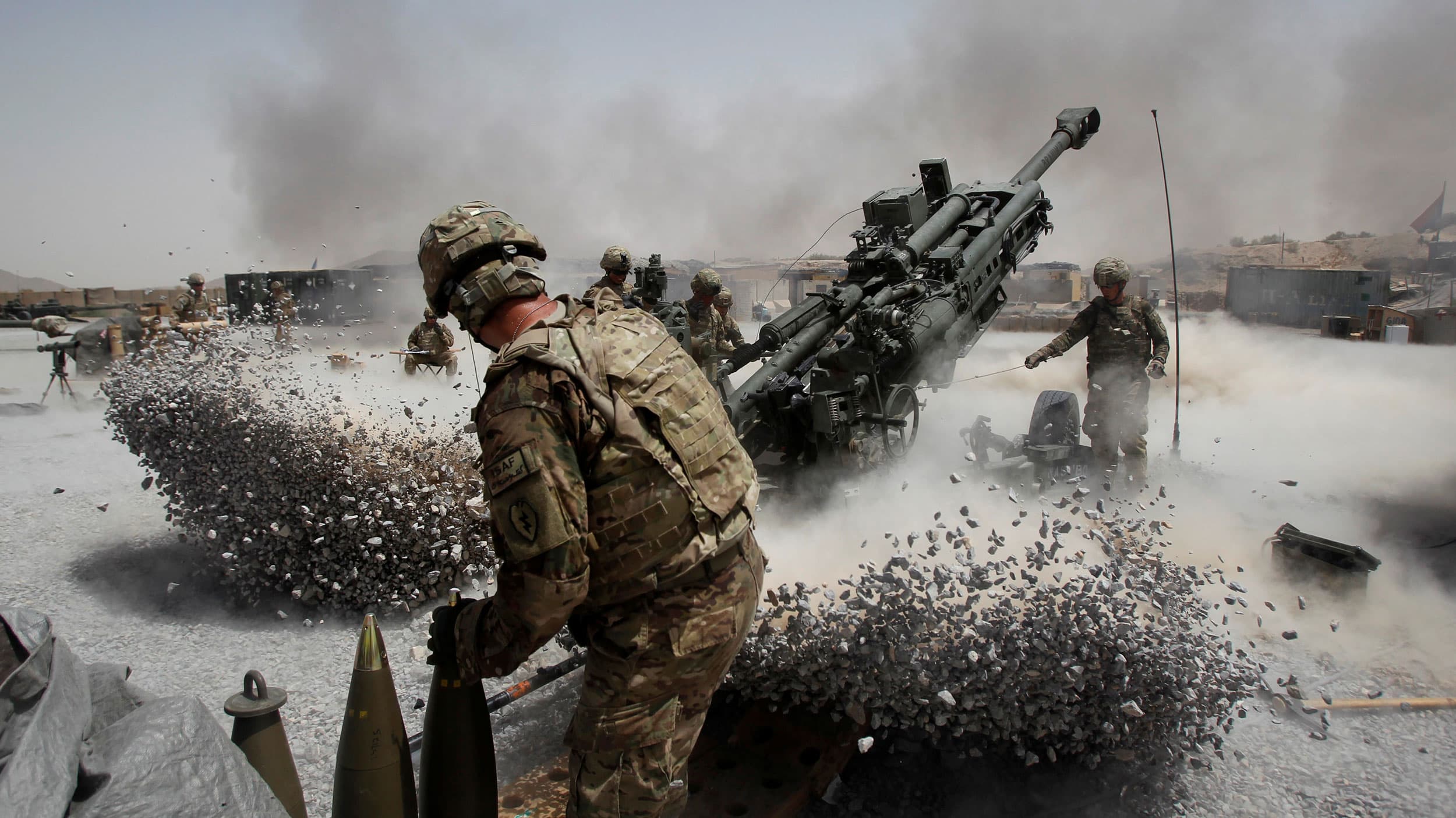US Army soldier is show firing a howitzer weapon, the exhaust of which has made piles of rocks fly on opposite sides of the artillary.