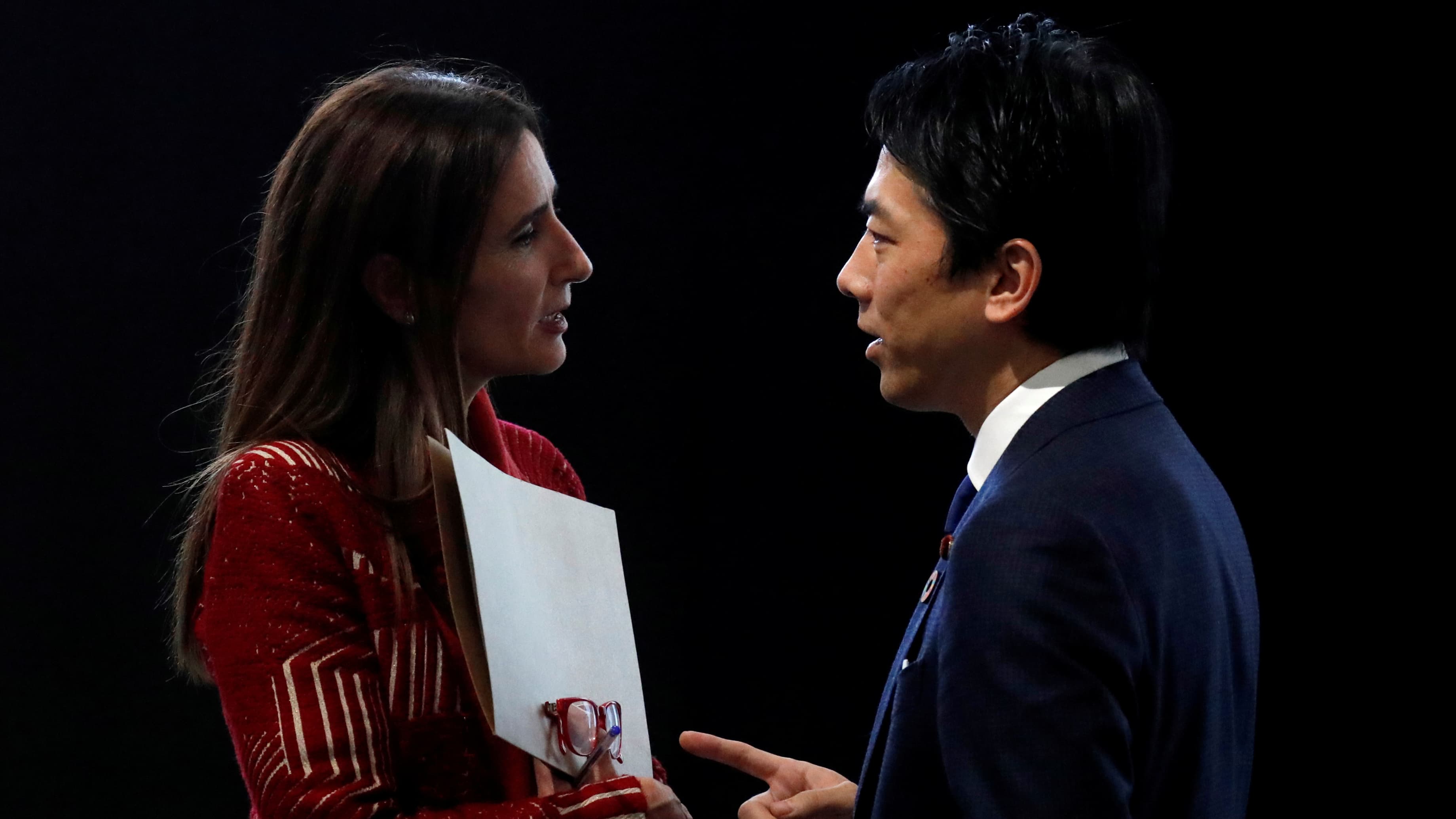 A woman speaks to a man against a black backdrop.