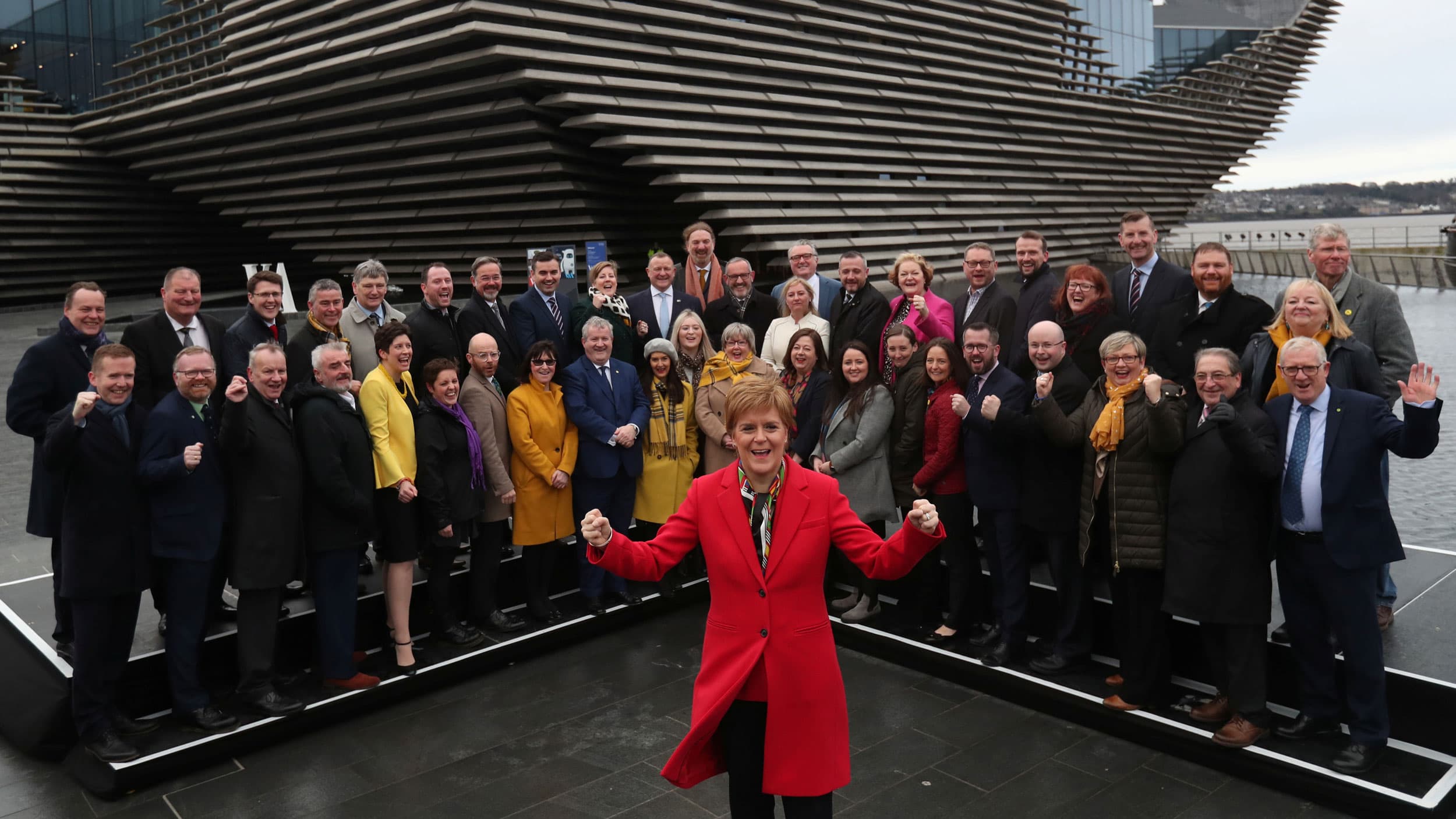 A woman in a red coat poses triumphantly in front of a group of happy adults.