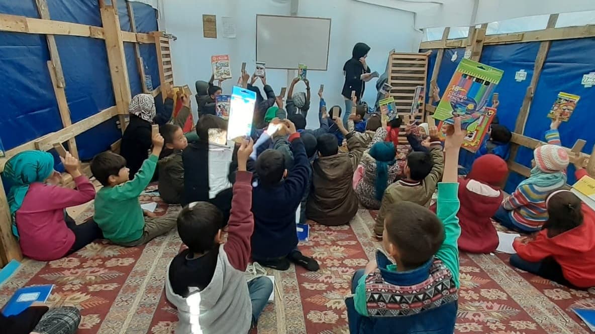A group of children sit in on carpet inside room surrounded by blue tarp