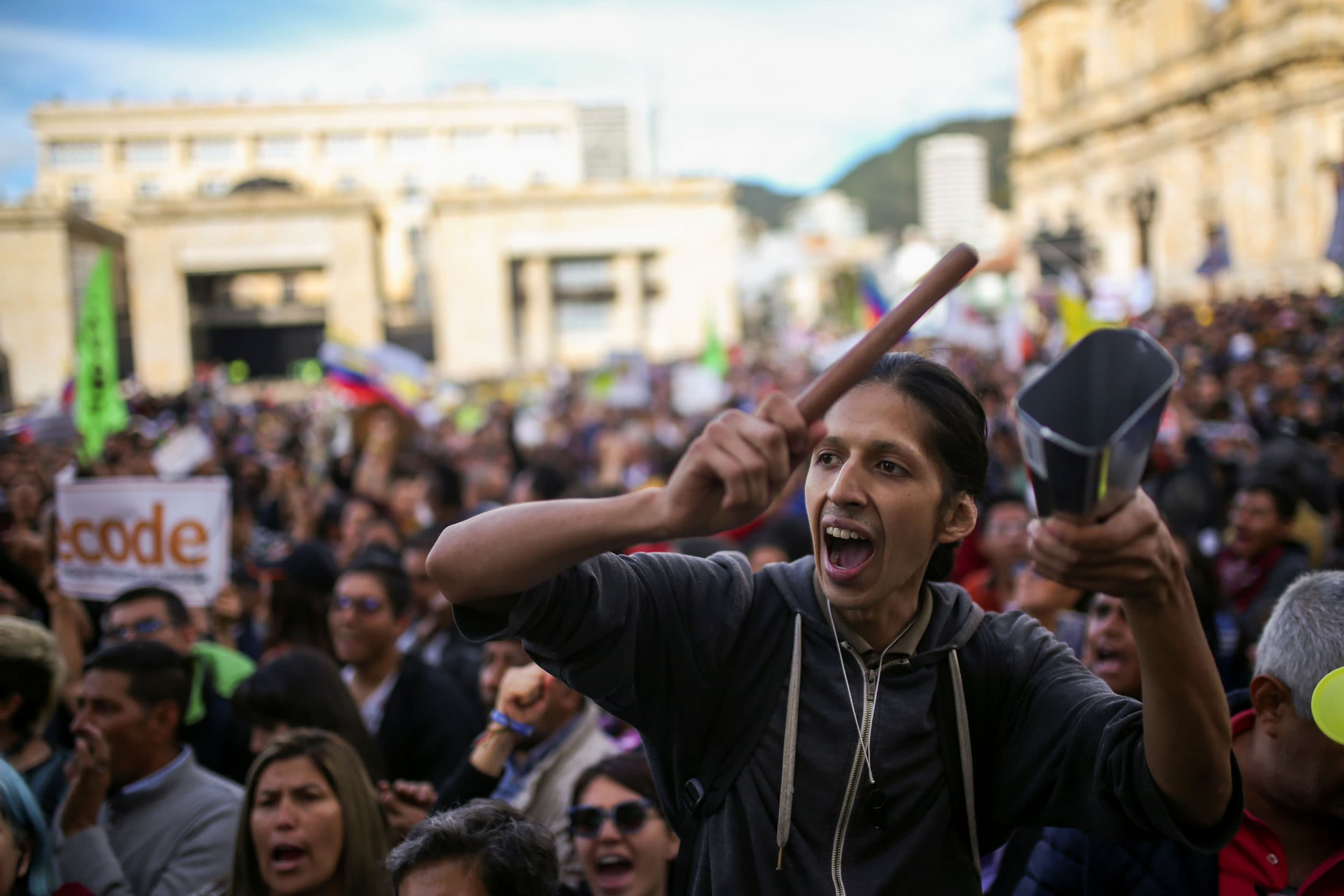 A large group of protesters are shown with a person in the nearground holding a cowbell.