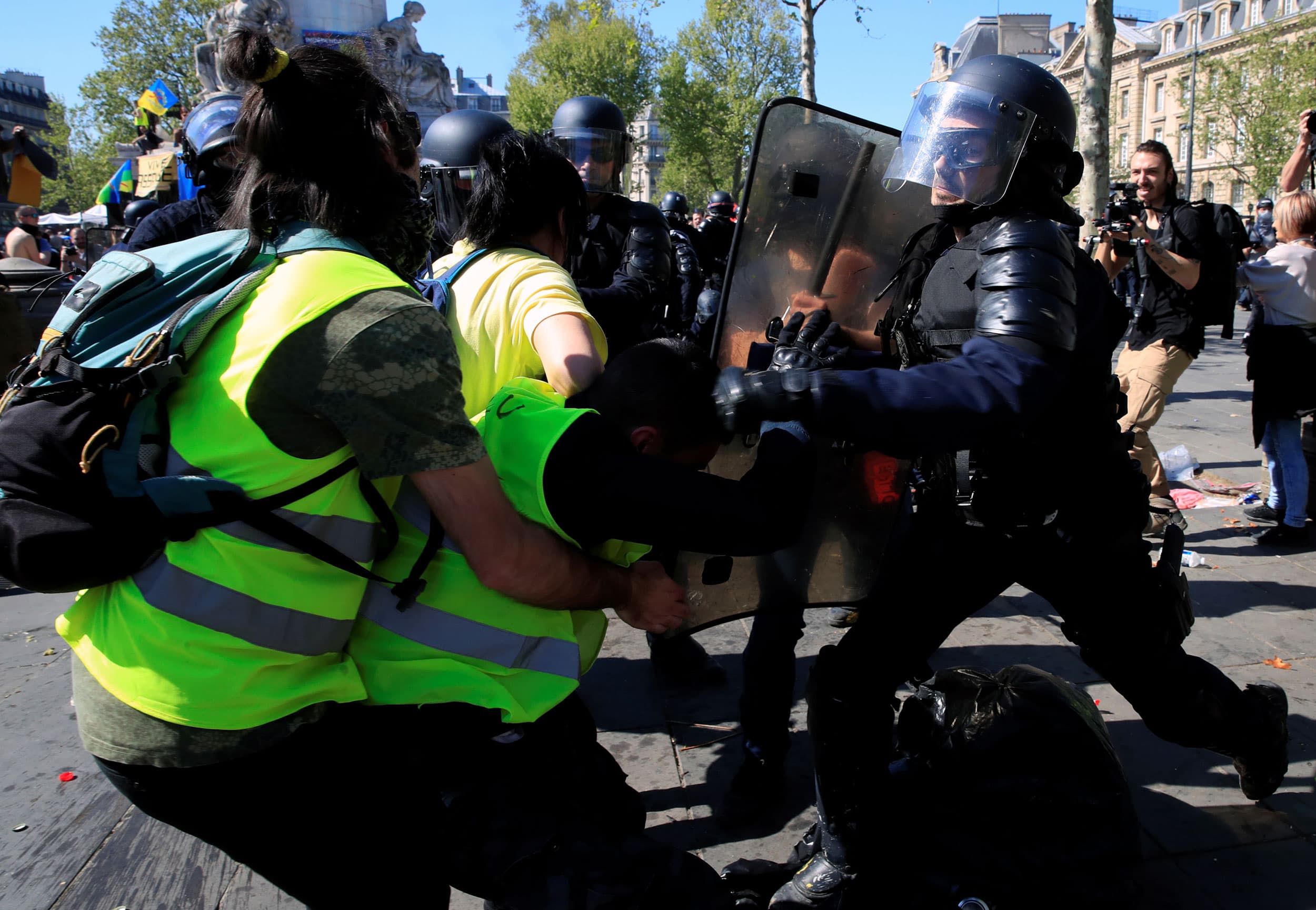 Two people wearing yellow safety vests are shown on one side with a police officer on the other carrying a shield.