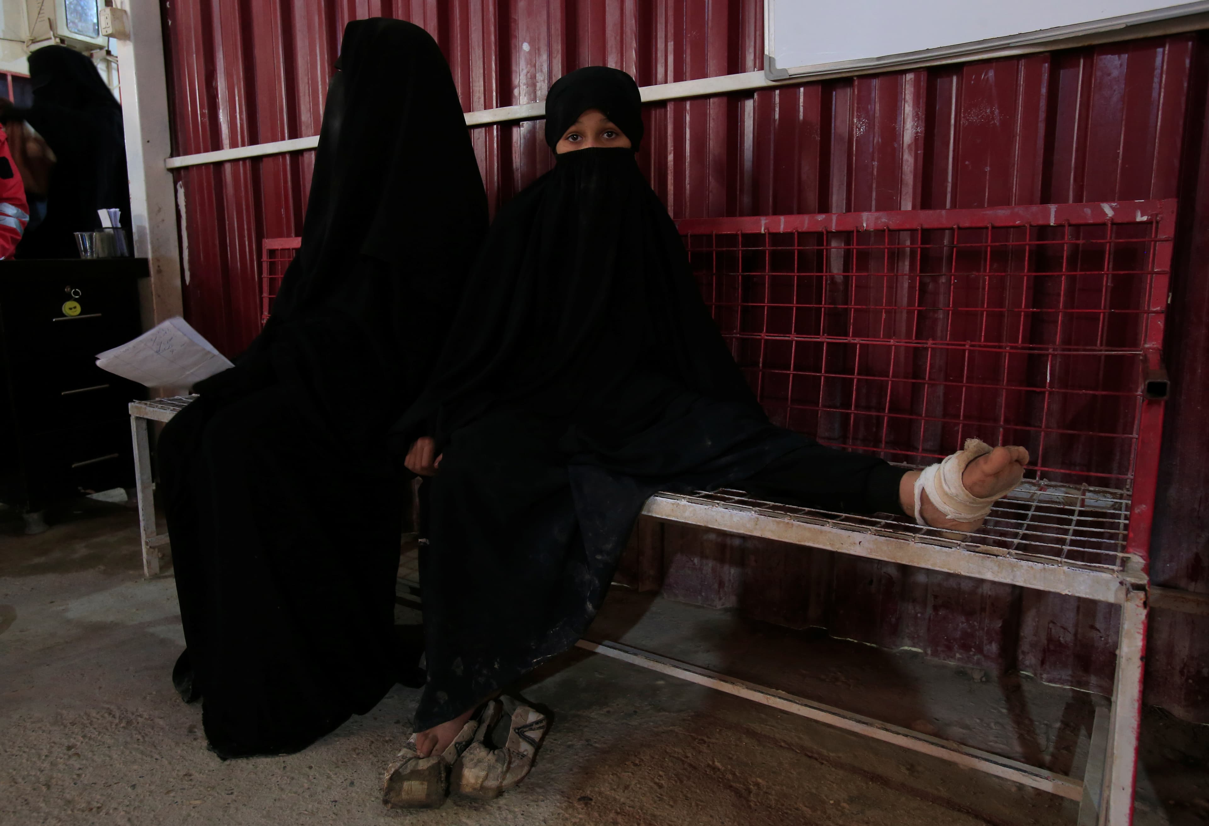 Two girls wearing black clothing sit on a bench in a clinic, one has a wounded foot wrapped in bandage