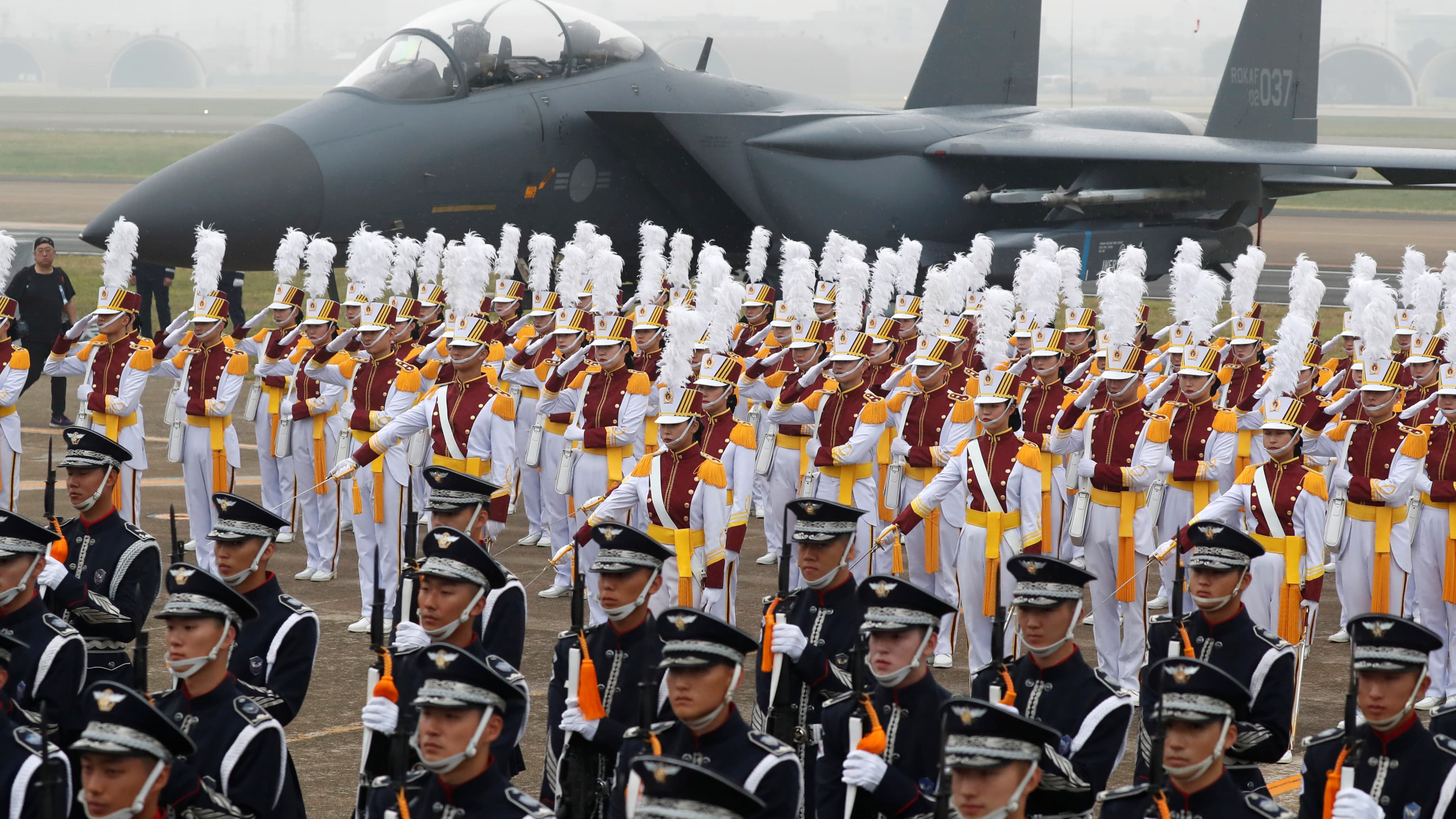 Rows of military men in white and black uniforms line up perfectly in front of a gray airplane