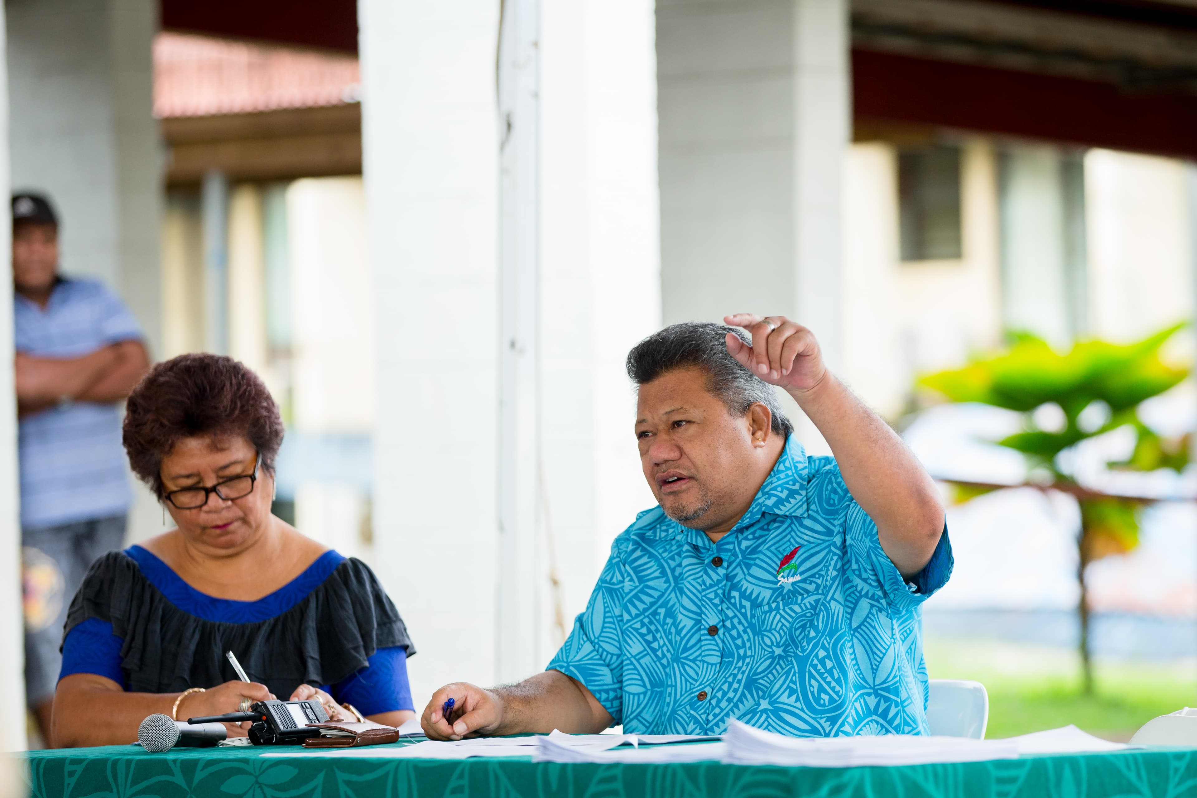 A man wearing a blue shirt speaks and points his finger in the air, sits next to a woman