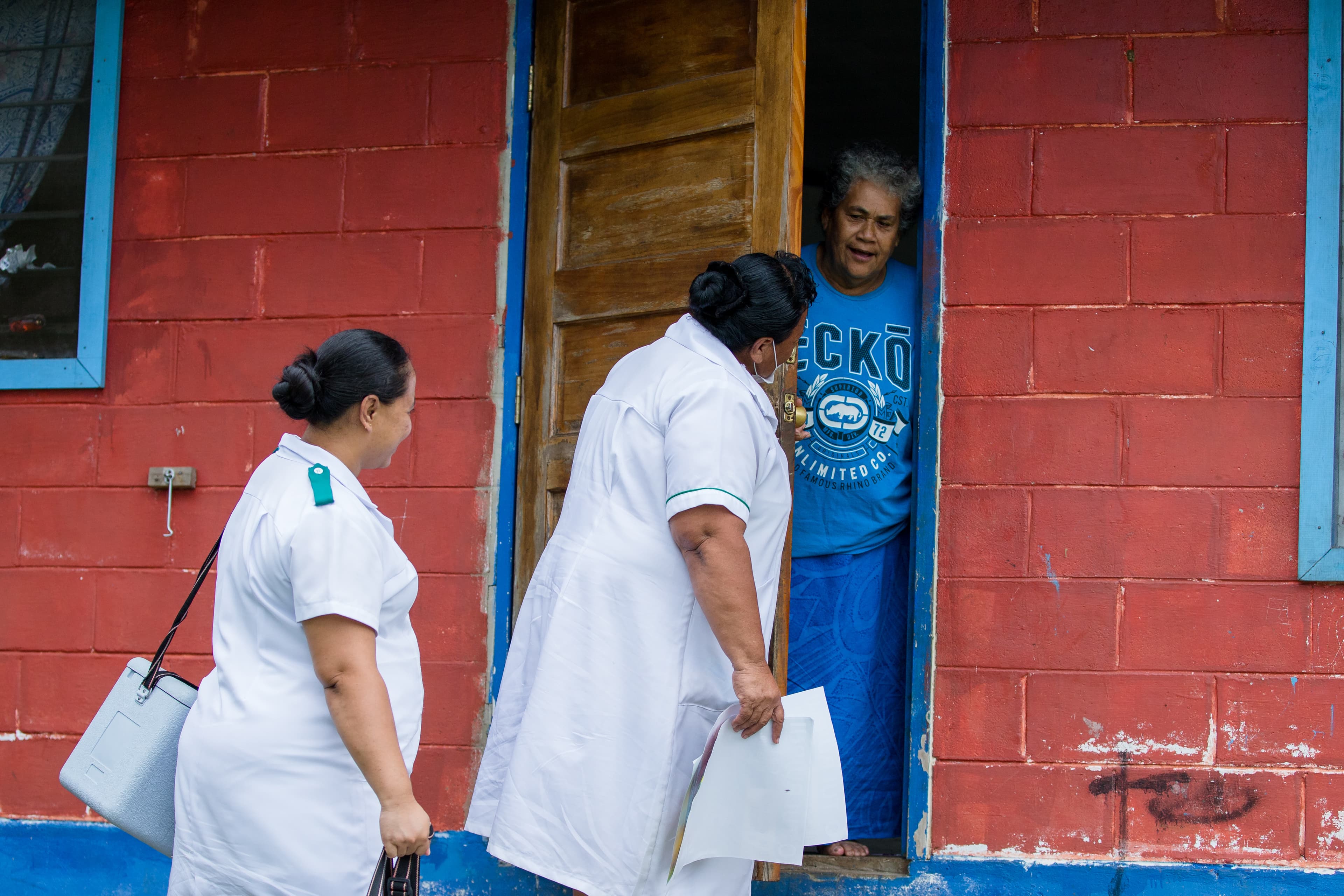 Two nurses stand in front of a house painted red and talk to a person standing at the door