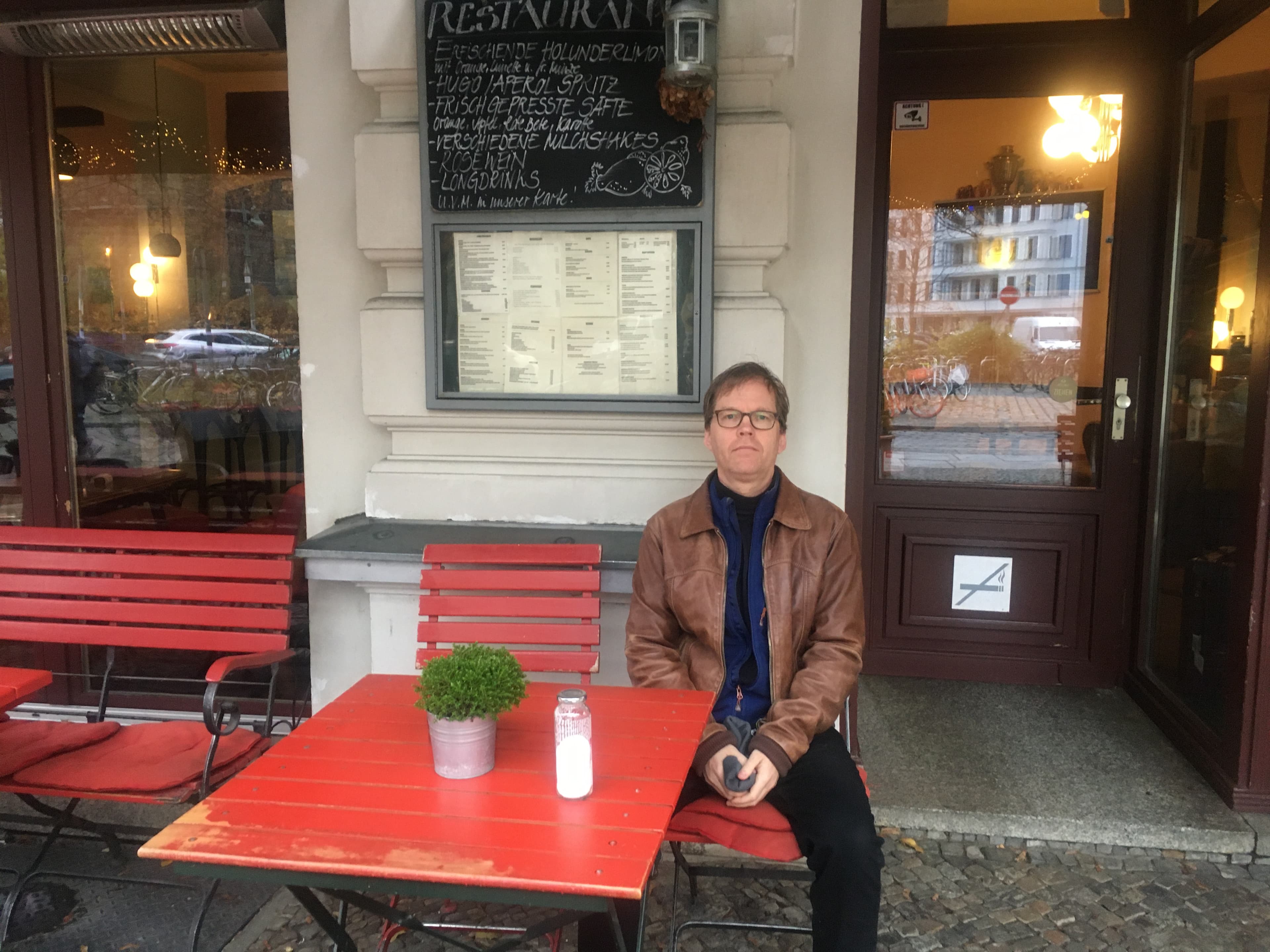 A man sits at an outdoor cafe with red tables and chairs