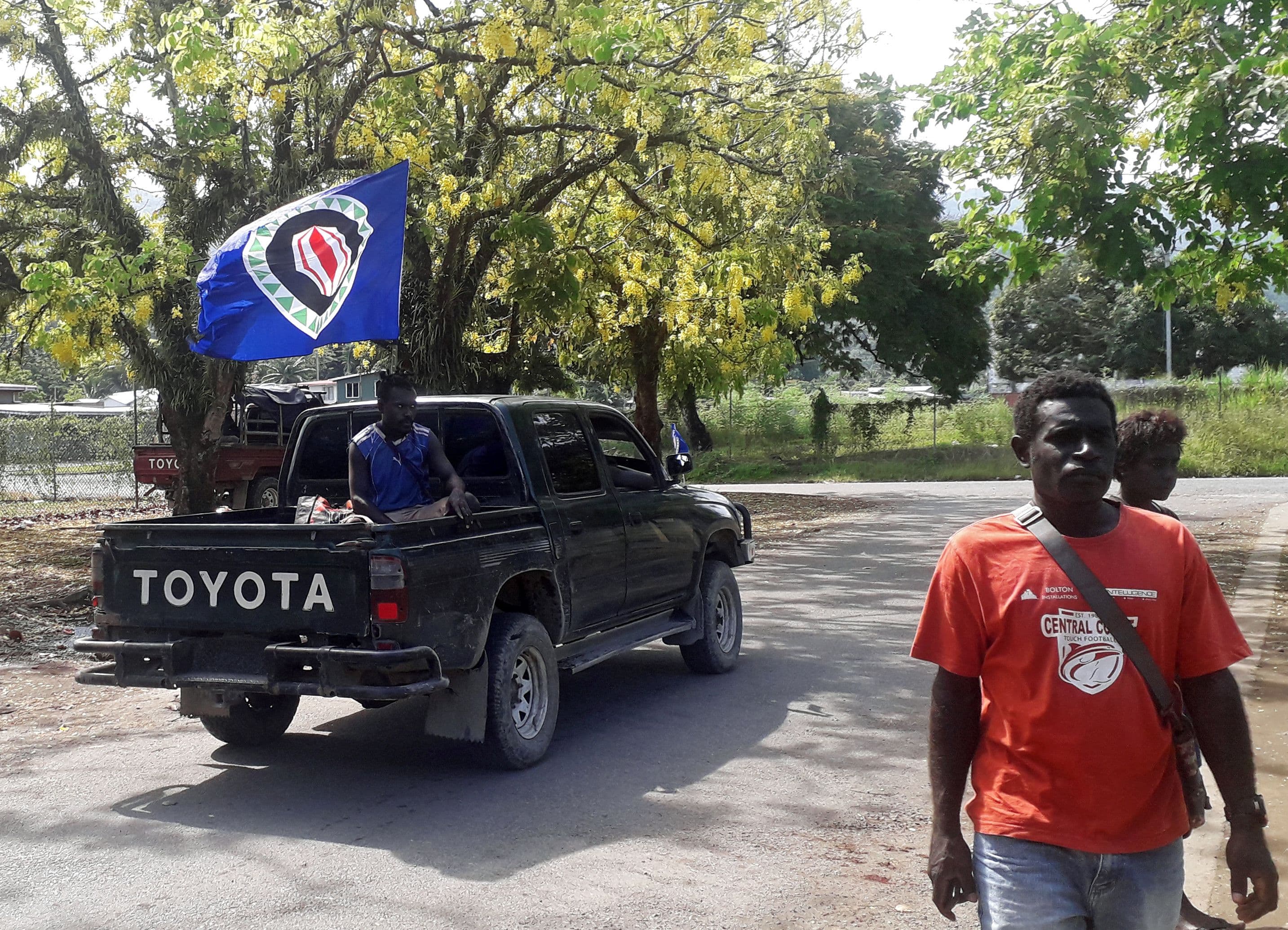 A truck carrying a Bougainville flag drives by a man.